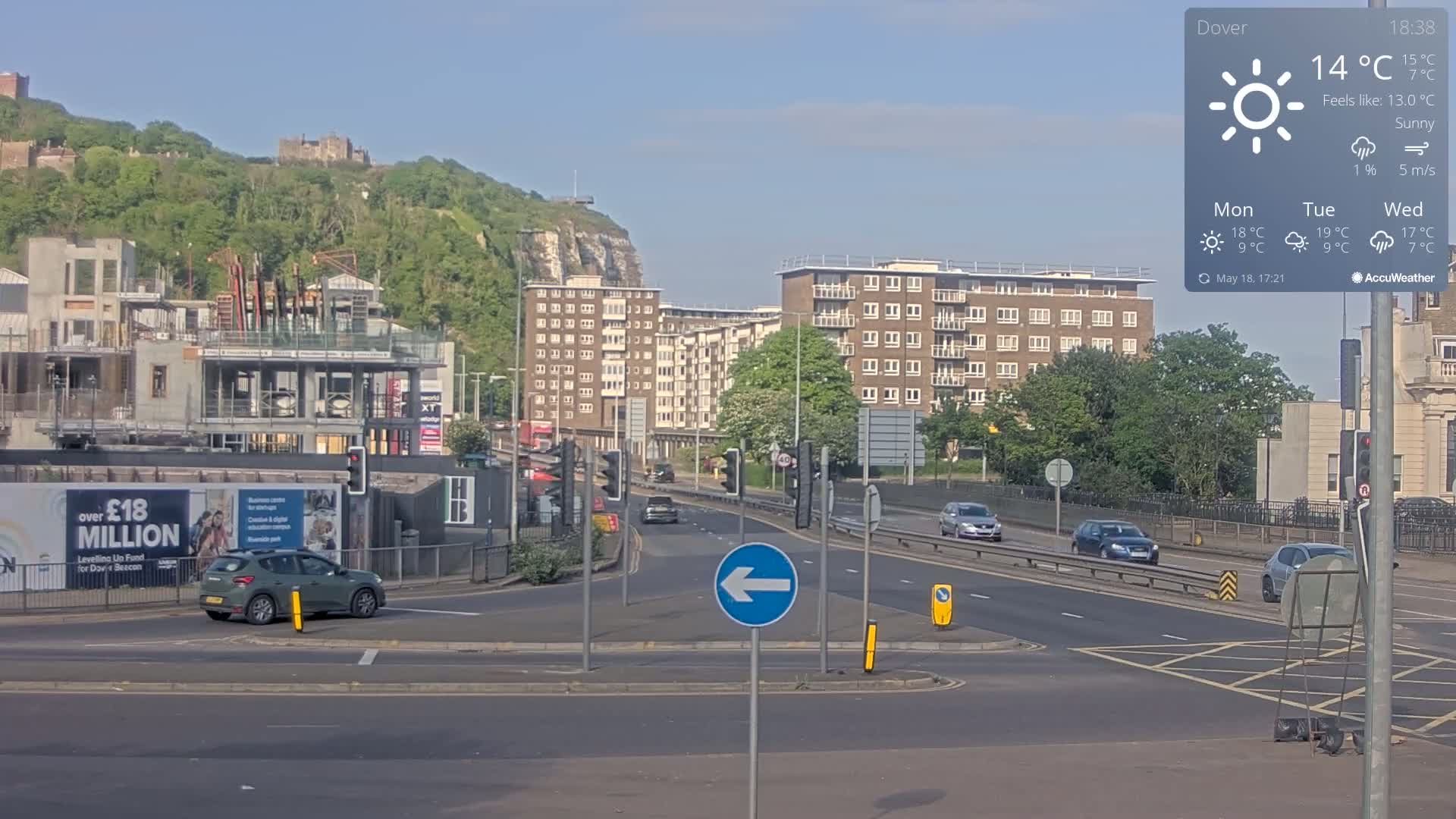 A sunny road intersection with several cars and buildings, including a partially constructed building, overlooks a hill with a castle in the distance.