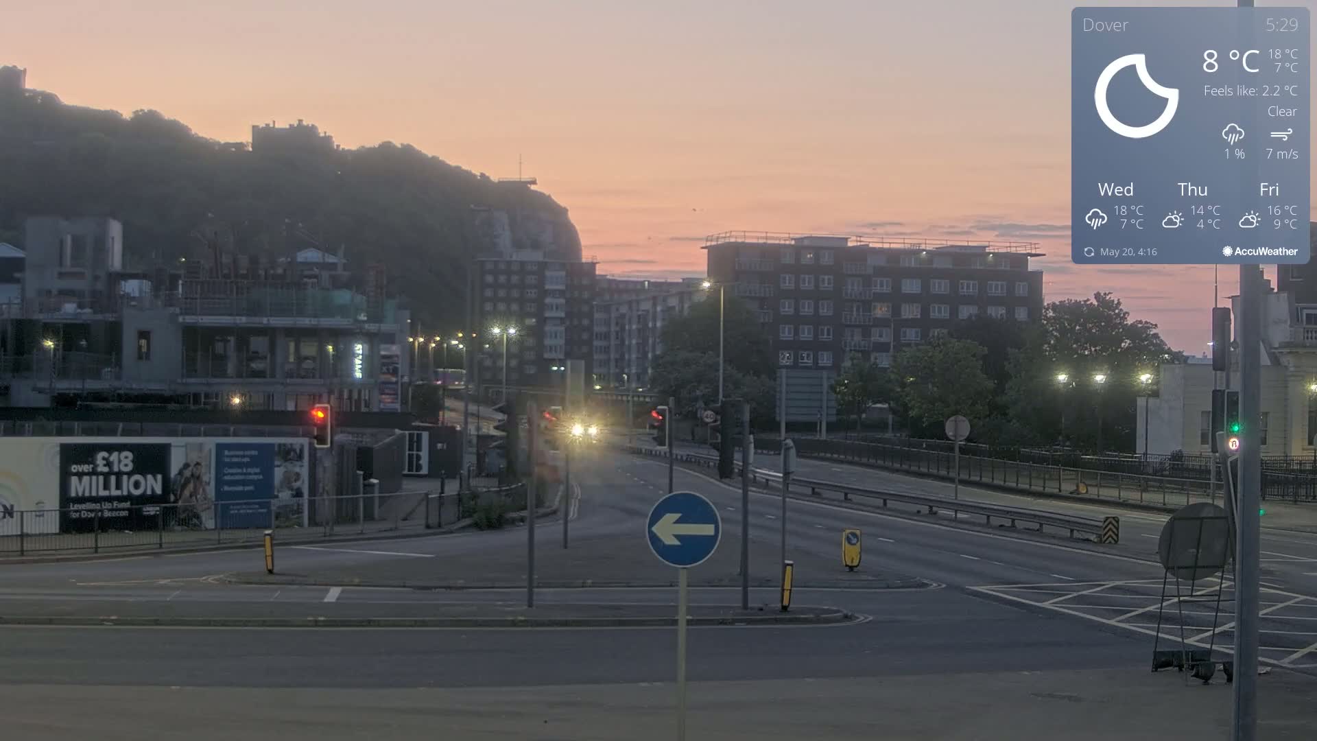 A mostly clear, dusky pink sunrise illuminates a city intersection with buildings, a hill in the background, and a few cars on the road.