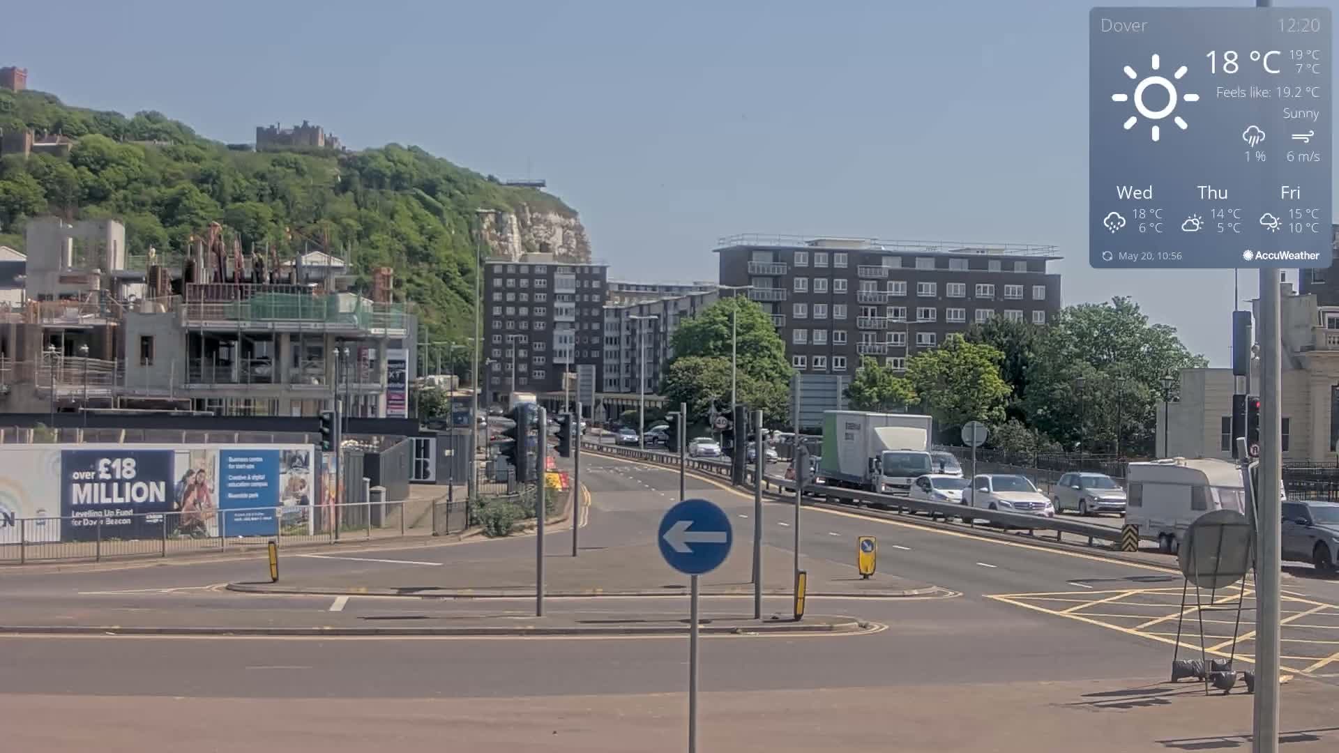 A sunny day reveals a cityscape with ongoing construction, apartment buildings, and moderate traffic on a roadway below a hill featuring greenery and a castle.
