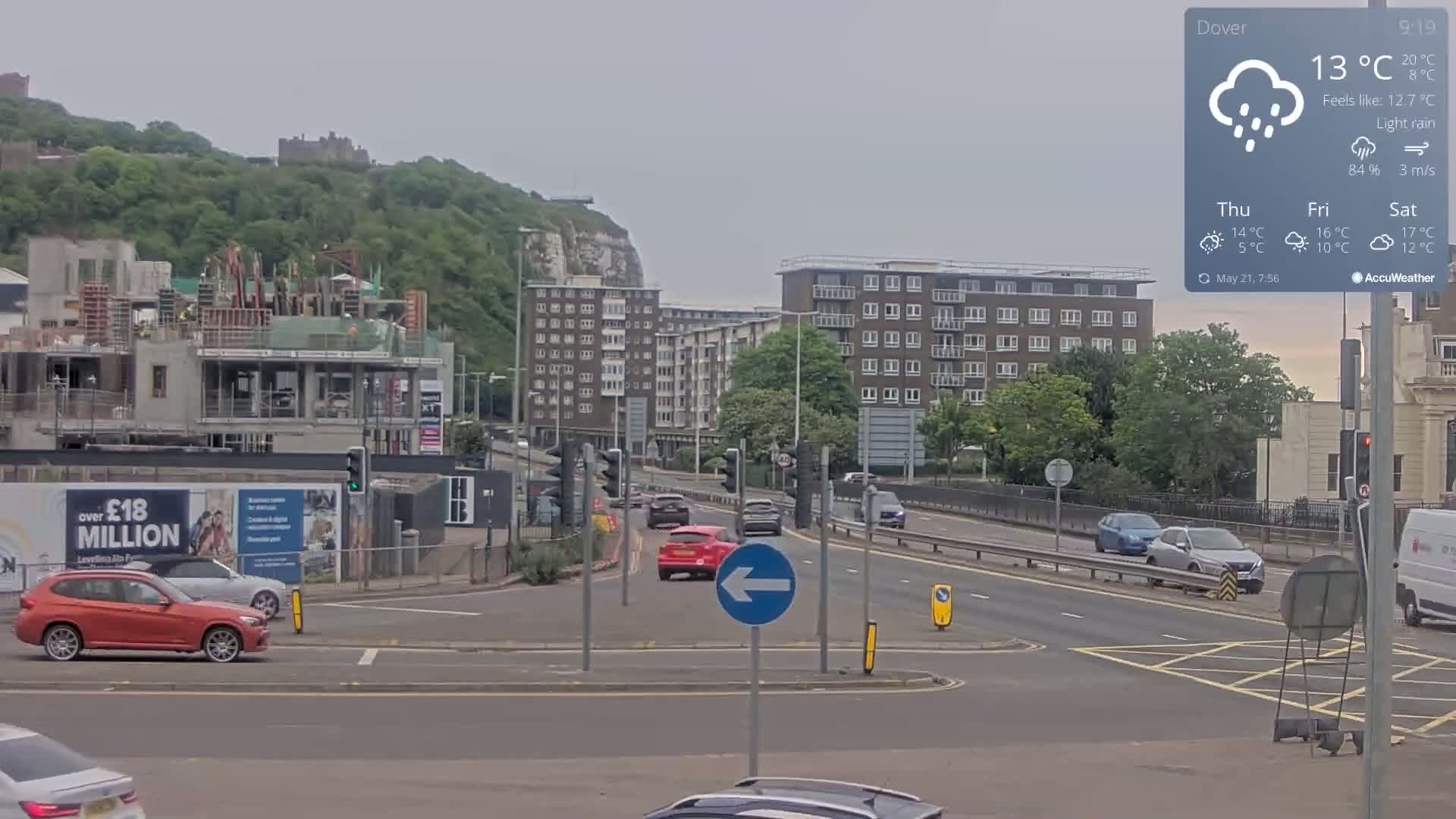 A roundabout with light traffic under light rain, near a partially-constructed building, apartment buildings, and a hill with a castle in the background.