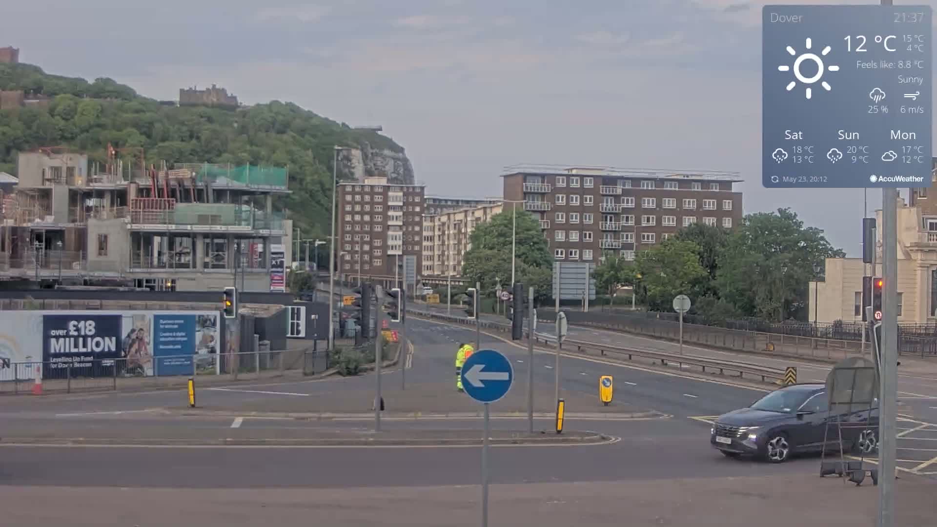 A sunny road scene shows a partially constructed building, apartment buildings, a hill with greenery and a castle in the distance, a dark-colored SUV, and a person in a yellow safety vest standing near a crosswalk.