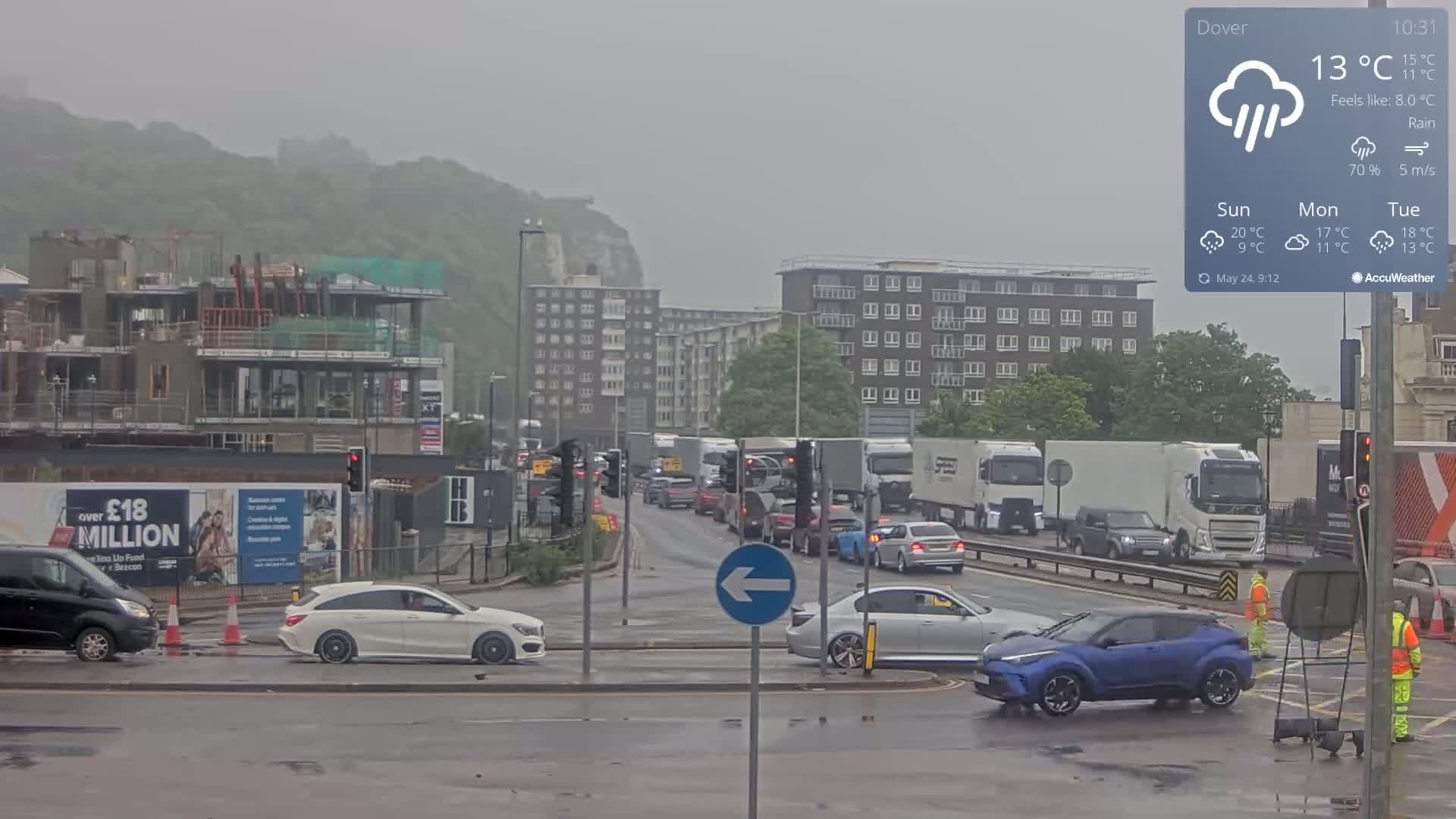 A rainy scene shows a busy intersection with several cars and trucks, a construction site, and apartment buildings, all set against a backdrop of a hill.
