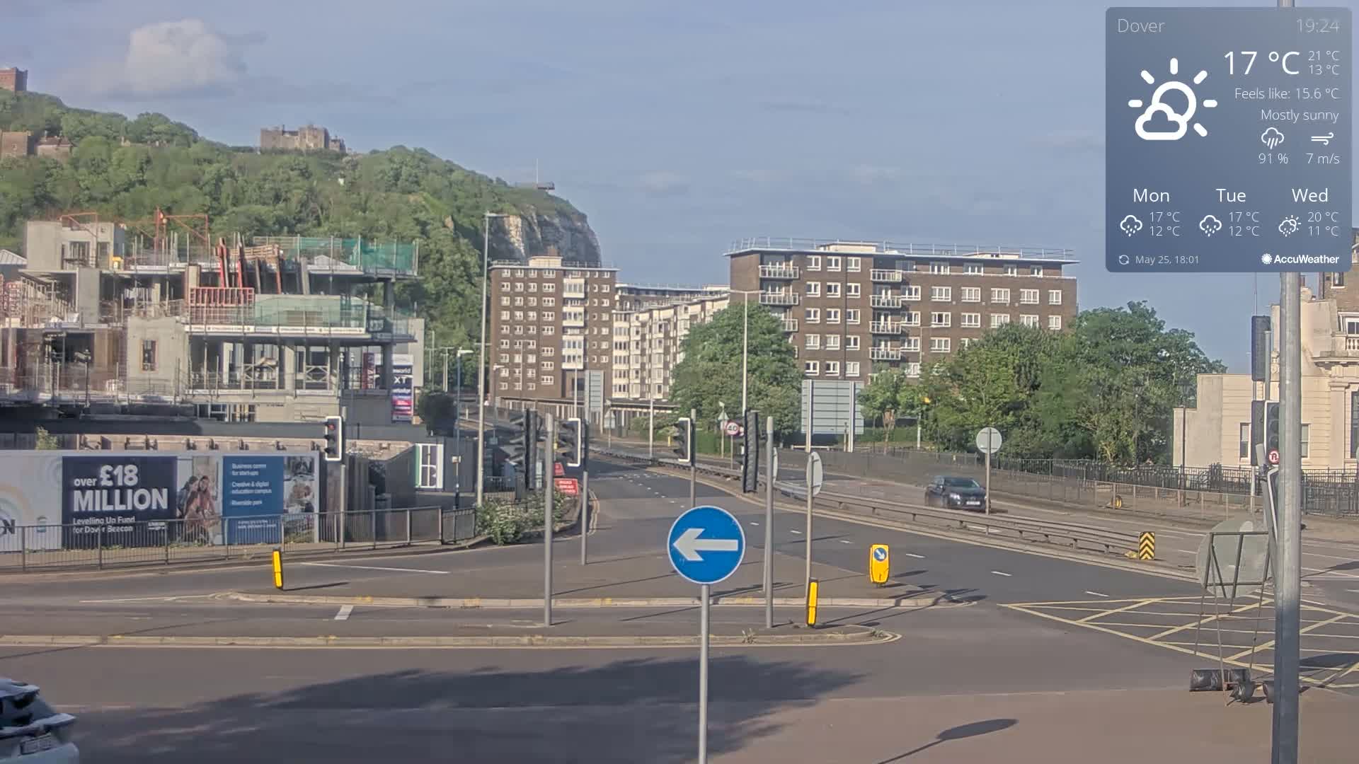 A mostly sunny road scene shows a multi-lane road with a roundabout, construction of a large building, apartment buildings, and a hill with a castle in the background.