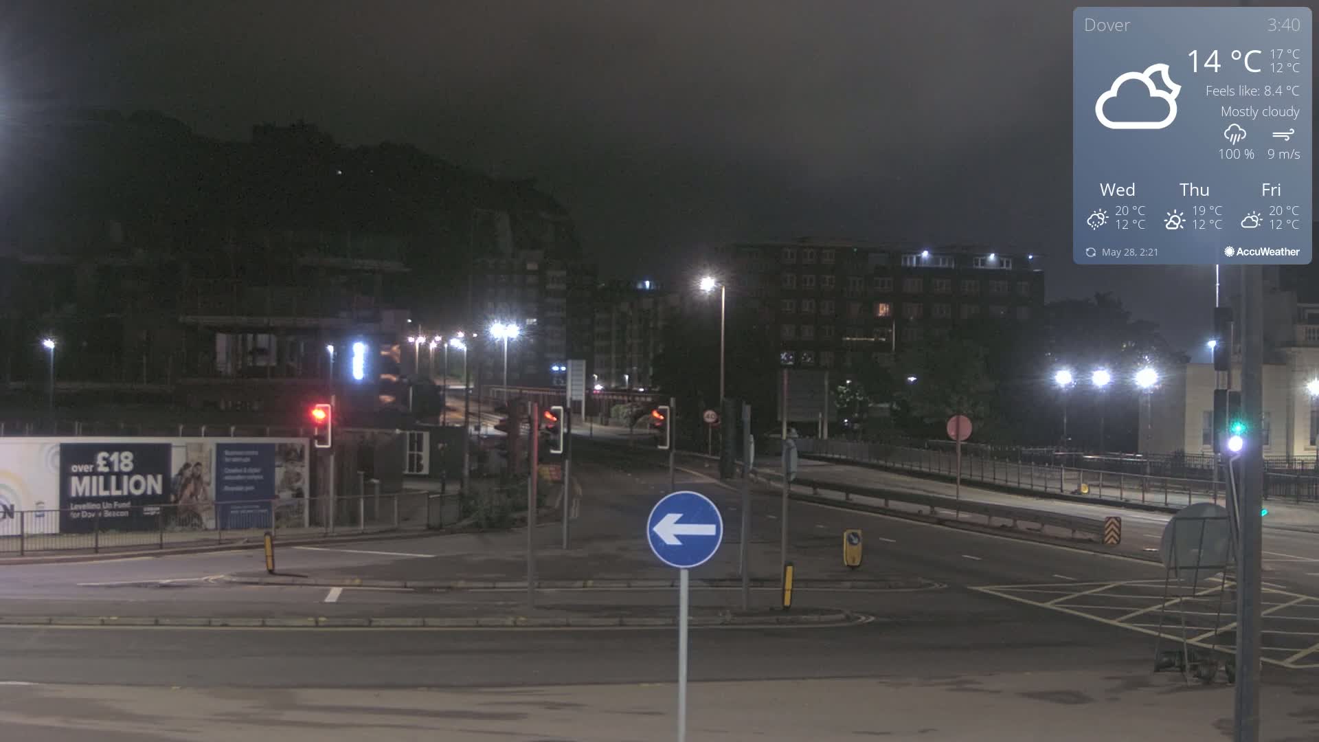 A mostly cloudy night scene shows a mostly empty intersection with streetlights illuminating buildings and a construction site under a dark sky.