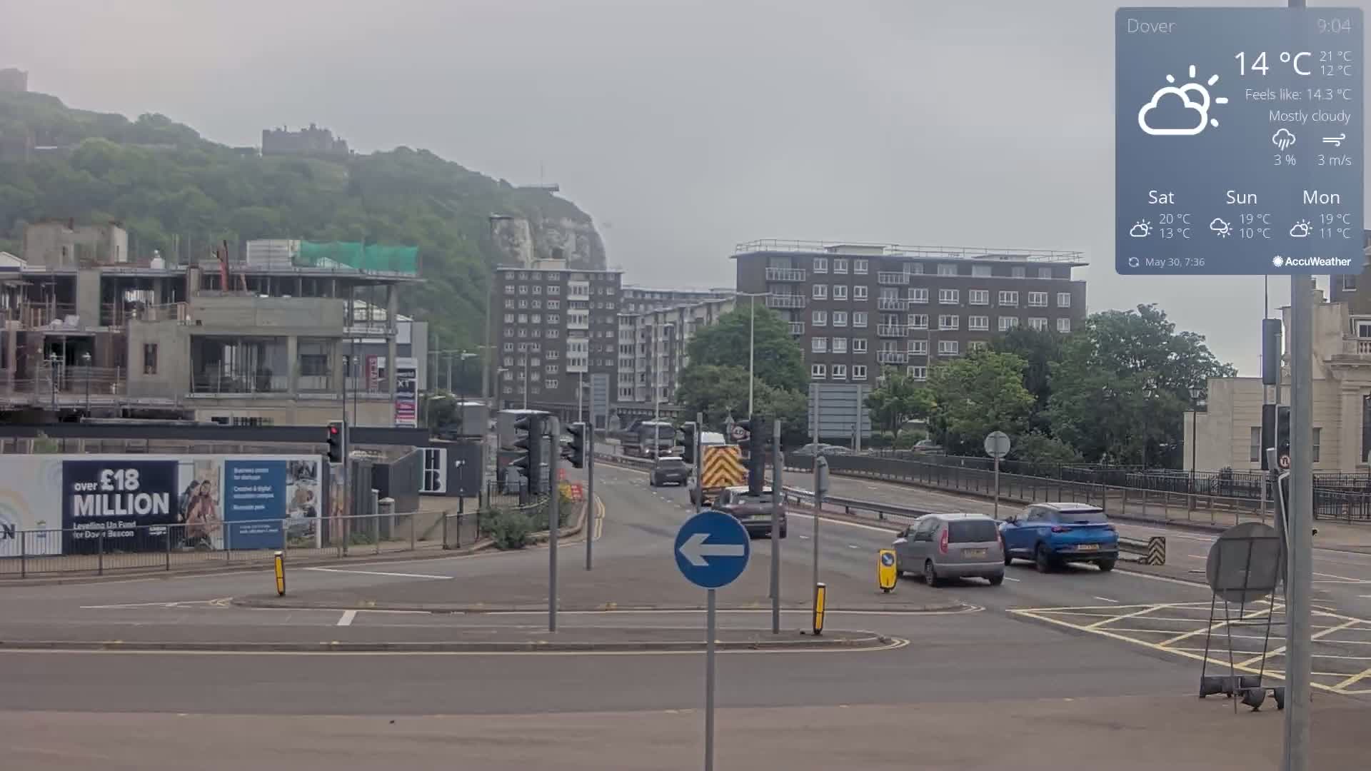 A mostly cloudy, overcast day shows a roadway with several cars, a construction site, apartment buildings, and a hill with greenery and a castle in the distance.