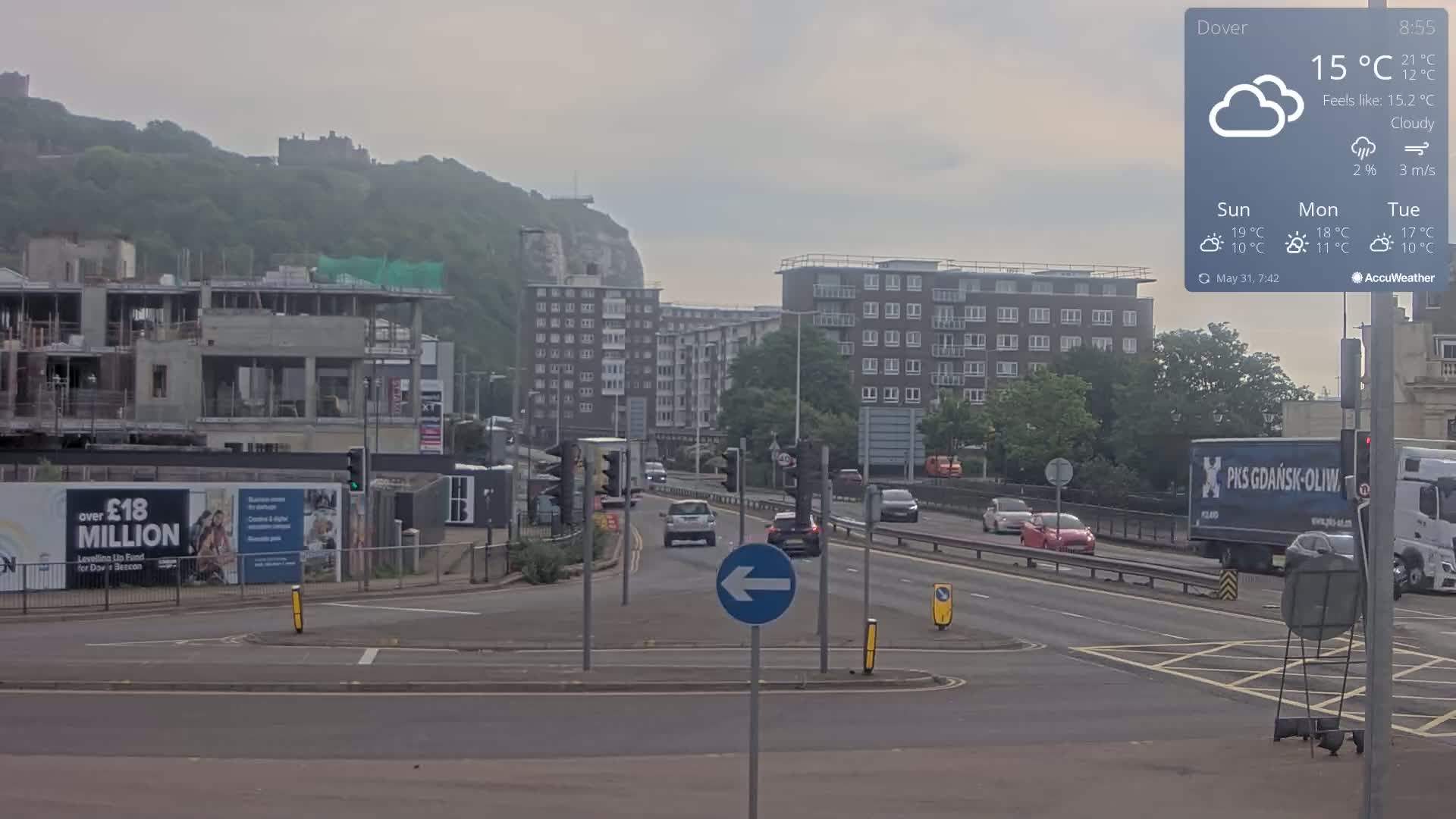 A cloudy day shows a multi-lane road with several cars and trucks, construction sites, and apartment buildings, all set against a backdrop of a hill with a castle.