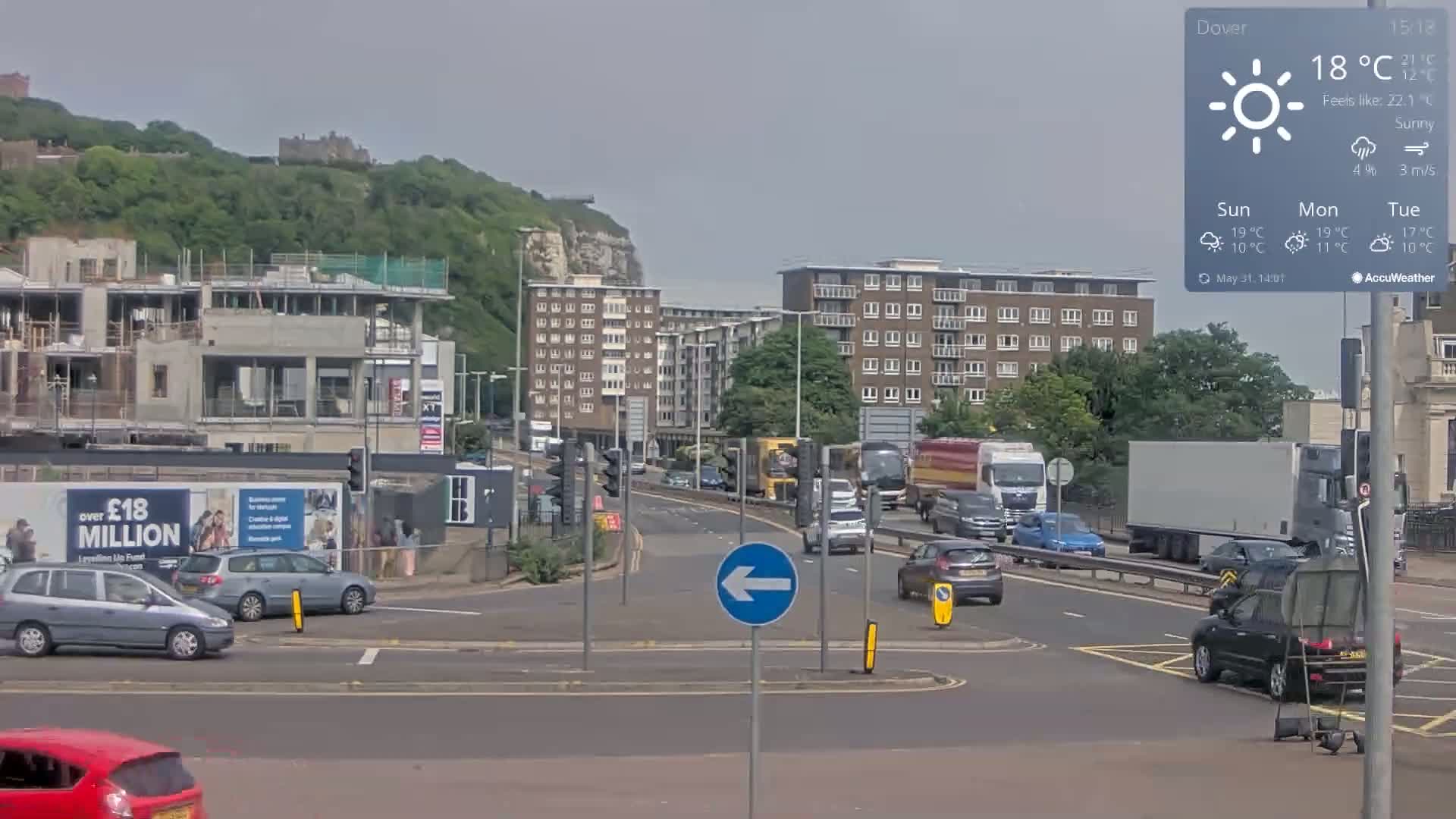 A sunny day shows a multi-lane road with cars and trucks, leading toward apartment buildings and a green hill with a building on top.