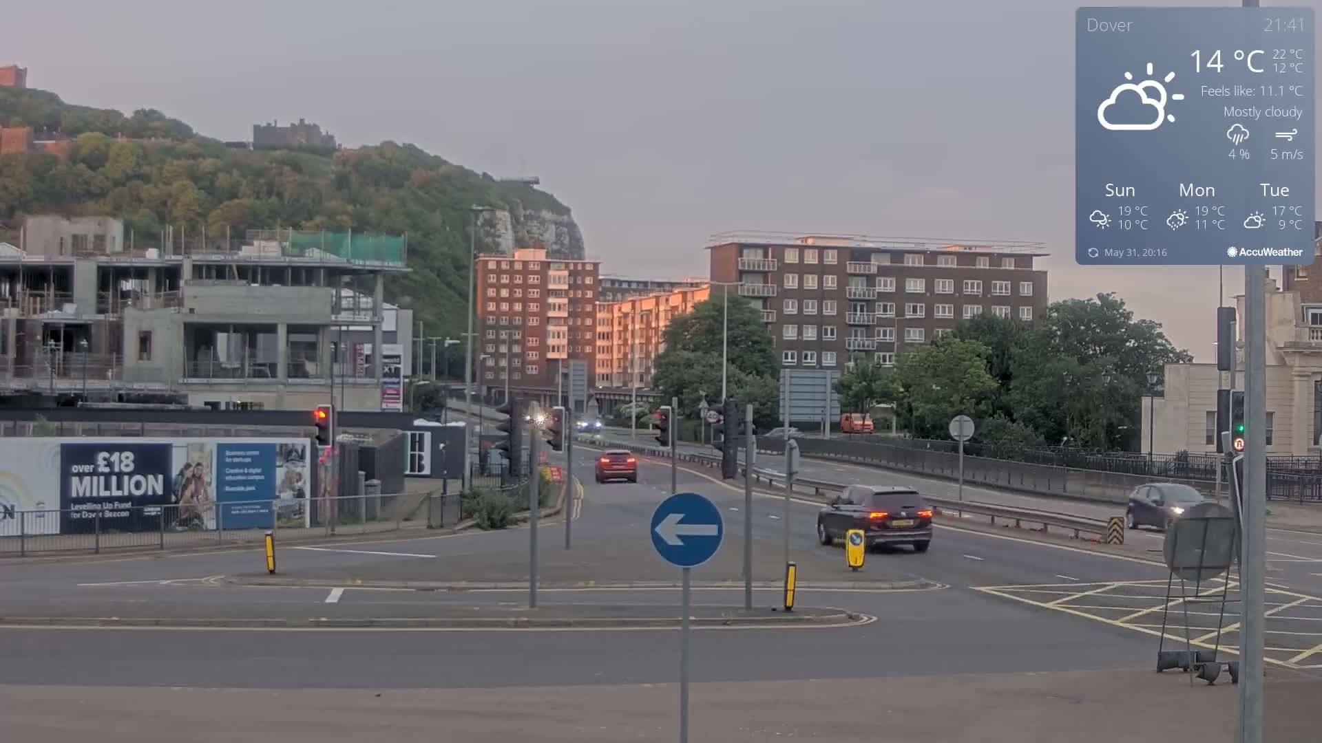 A mostly cloudy evening scene shows a multi-lane road intersection with several cars, a construction site, apartment buildings, and a castle on a hill in the background.