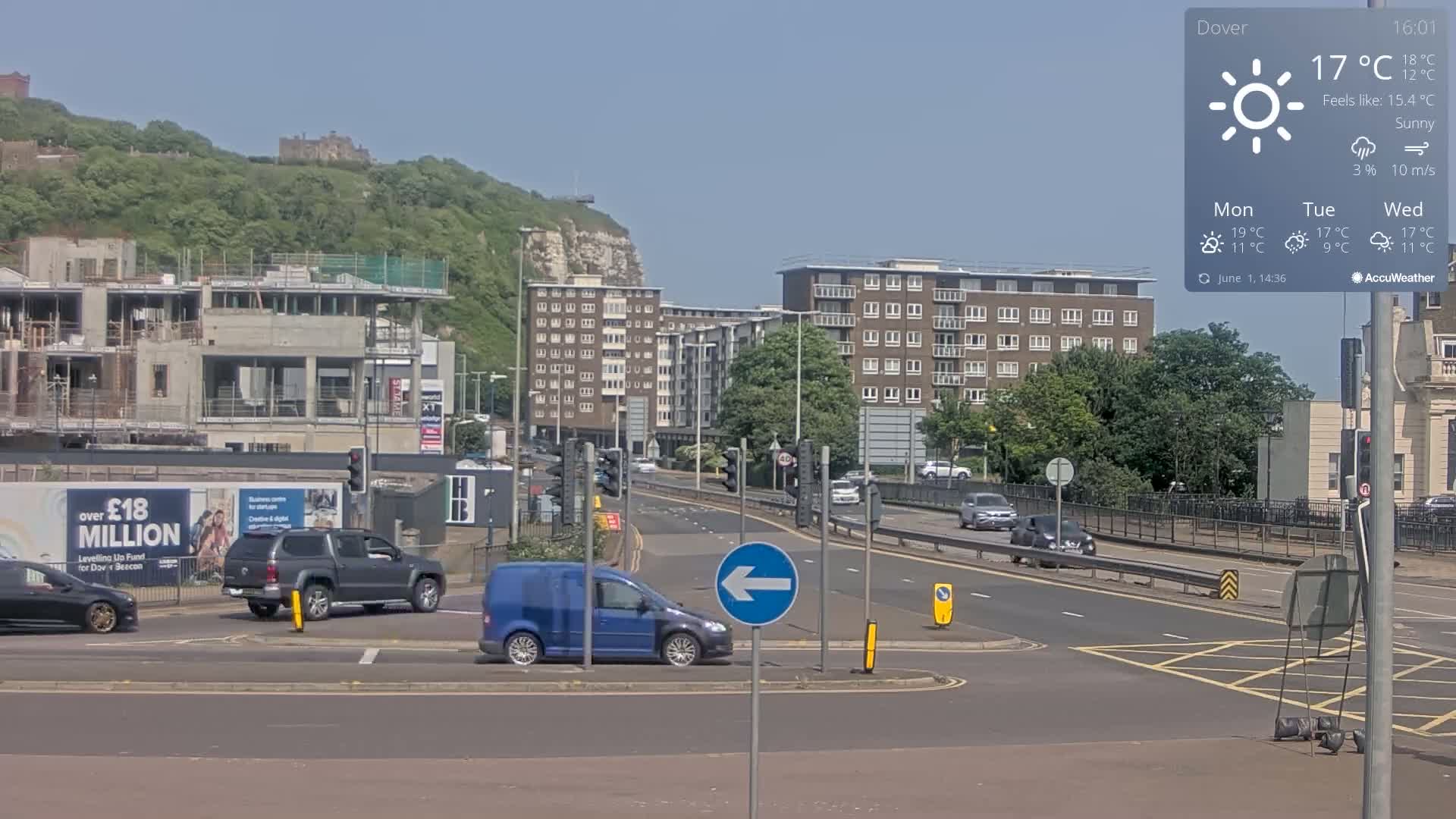 A sunny day shows a multi-lane road intersection with several cars, a blue van, and buildings, including apartment complexes and a construction site, with a hill and castle visible in the background.