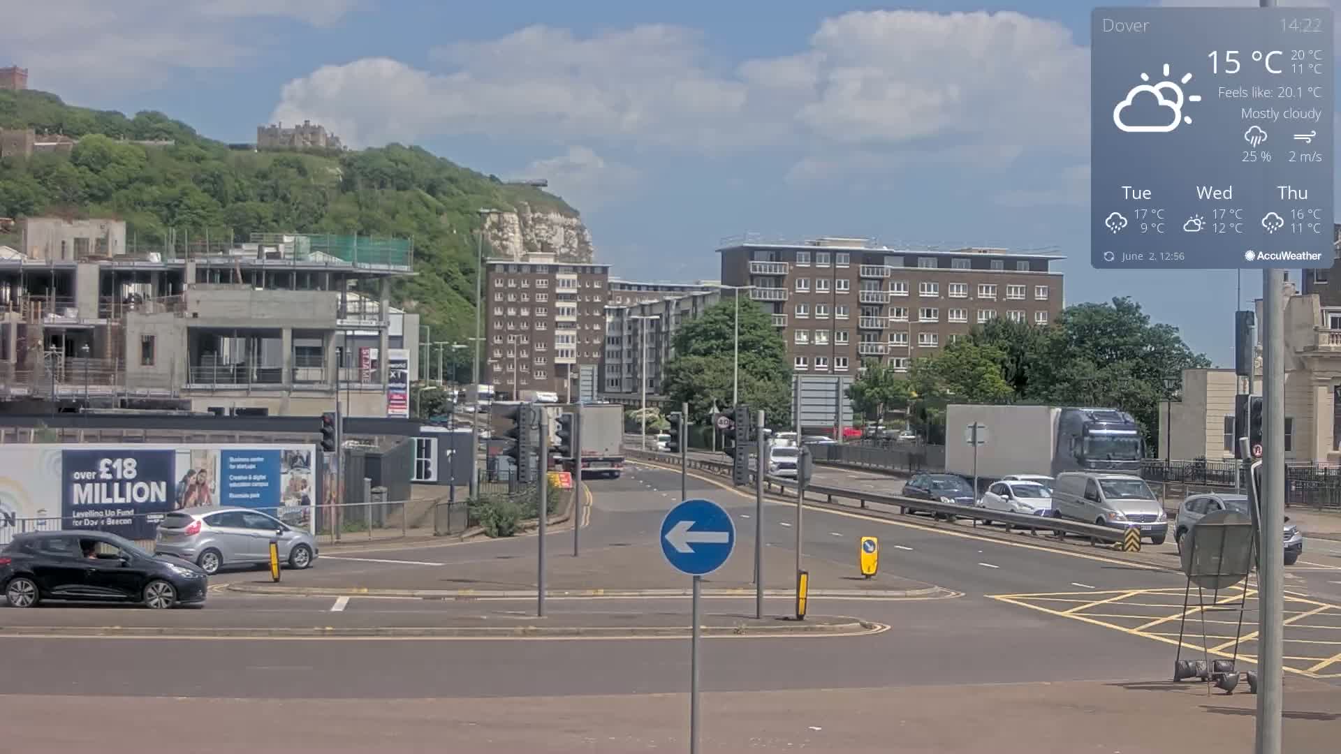 A mostly cloudy, sunny day shows a view of a city intersection with traffic, a construction site, apartment buildings, and a castle atop a distant hill.