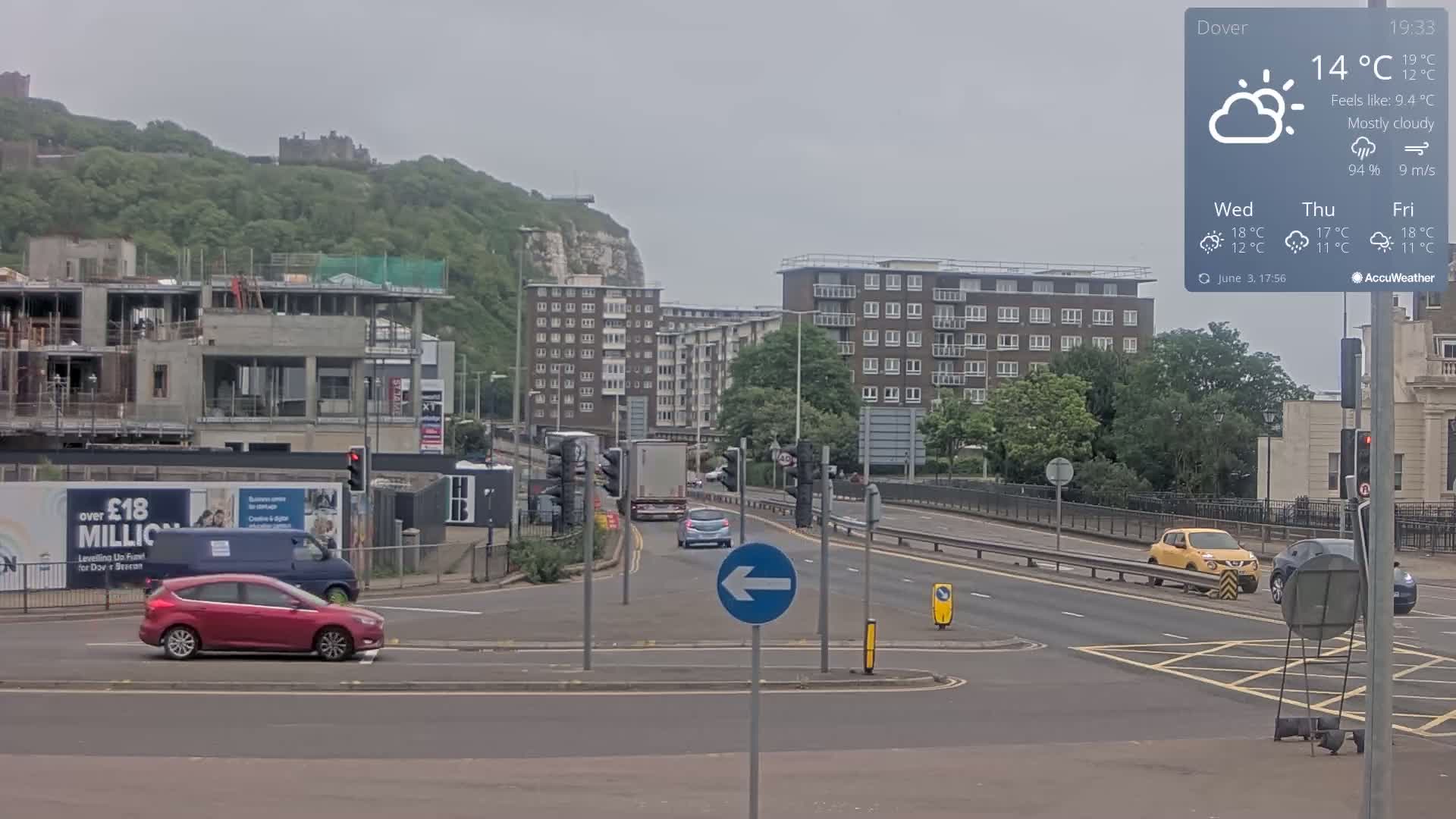 A mostly cloudy, 14°C day shows a highway intersection with several cars and buildings, including a partially constructed building and a hill with a castle in the distance.