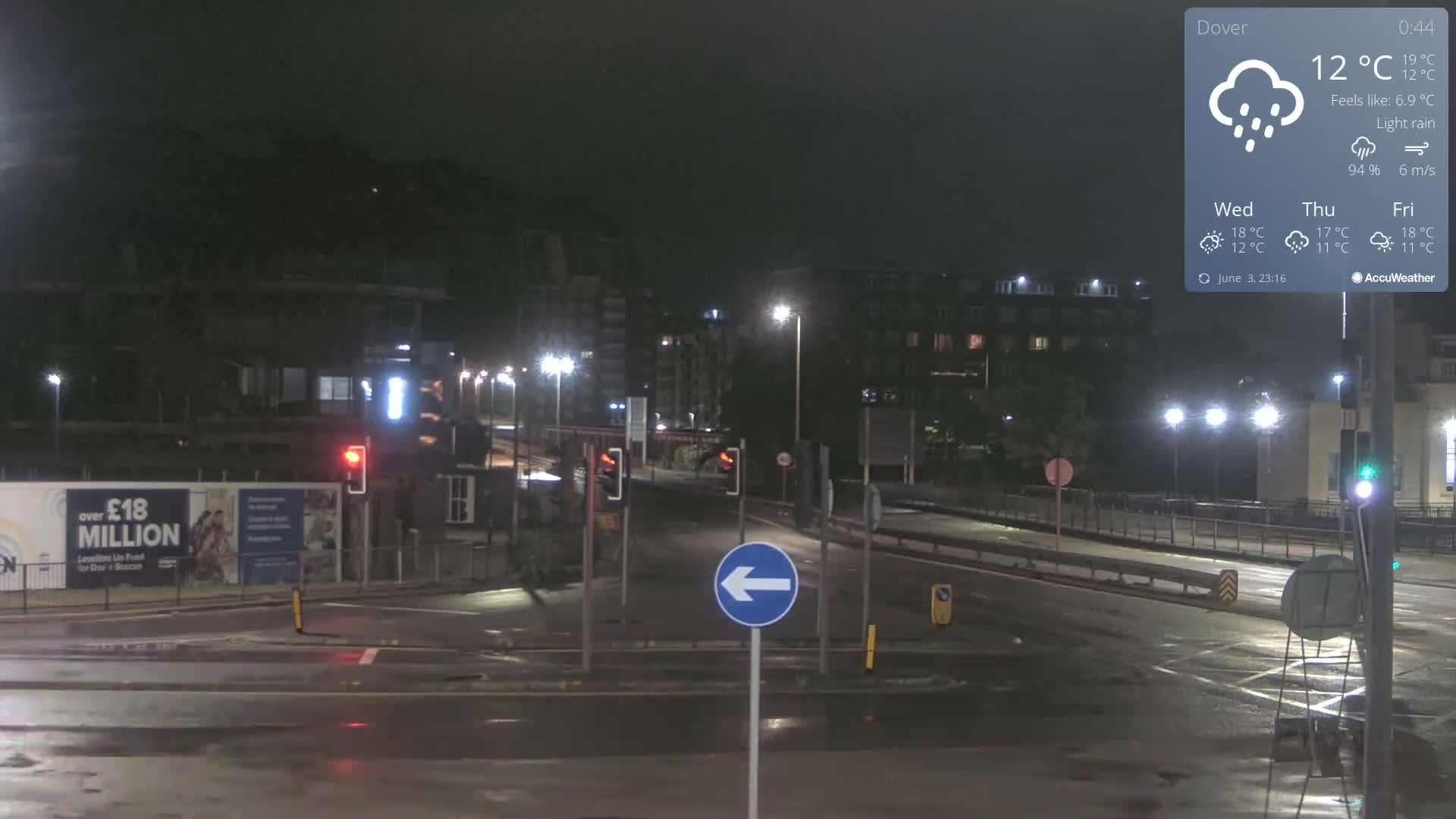 A nighttime, rain-slicked road intersection with streetlights and buildings in the background under light rain.