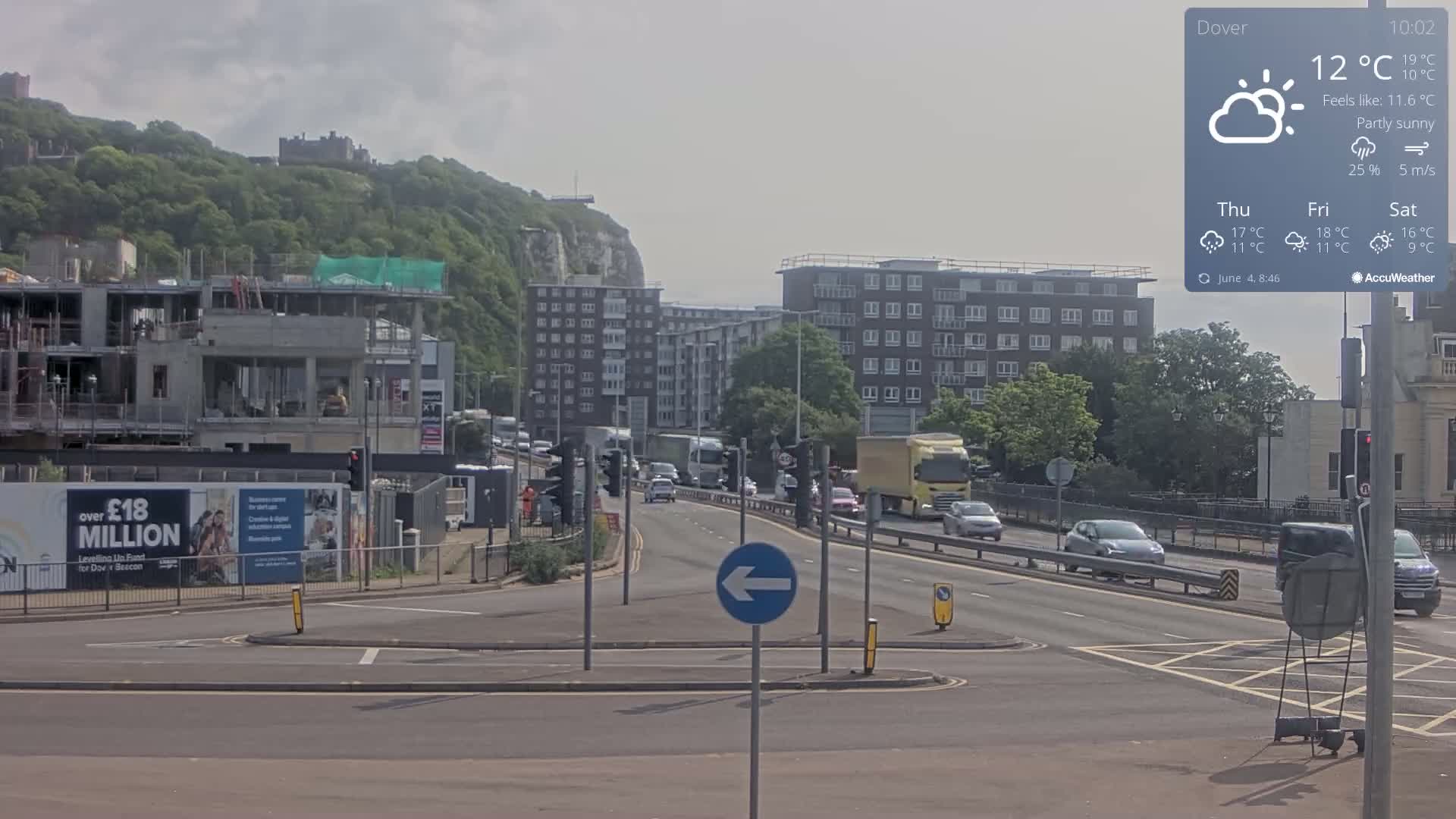 A partly sunny day shows a busy road intersection with cars and trucks, construction sites, and residential buildings, all situated below a hill with a castle visible in the distance.