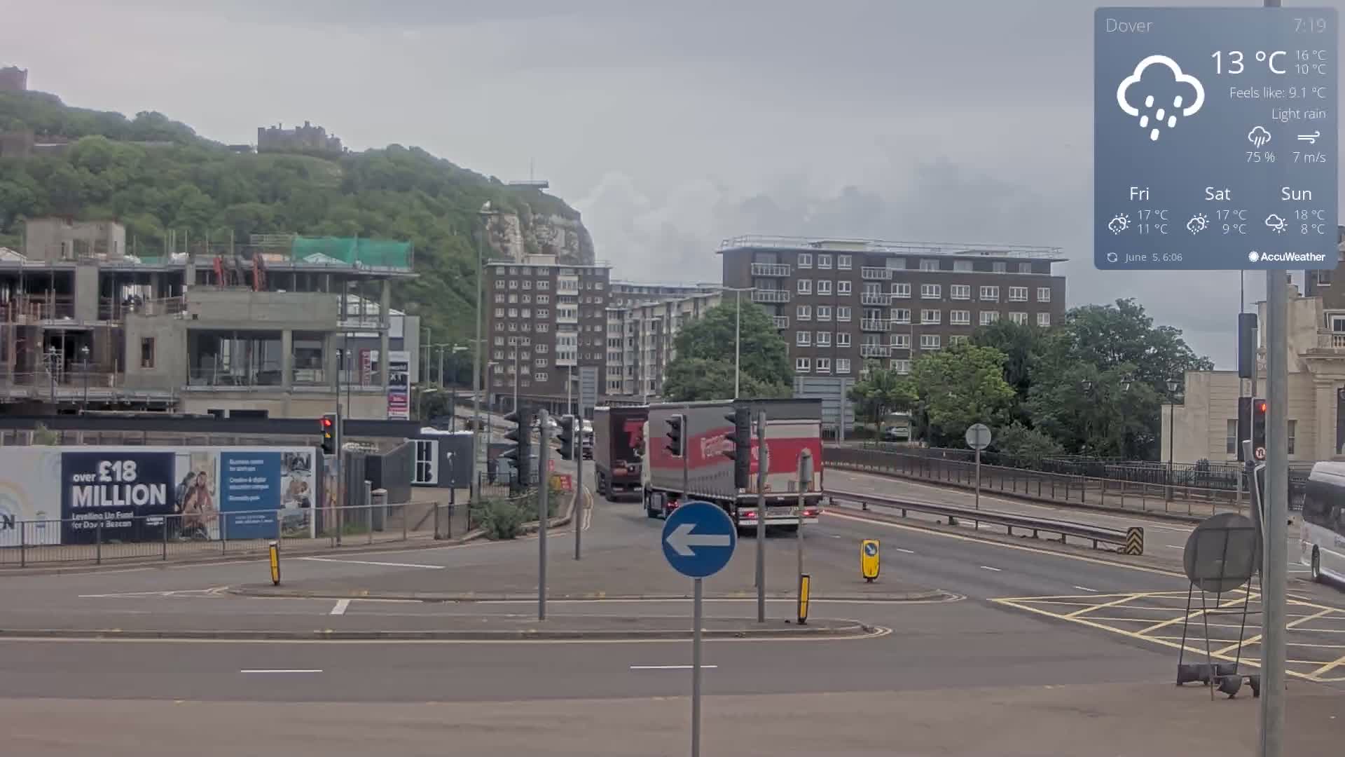 A city street scene under light rain shows a roundabout with several buildings, including a partially constructed one and apartment buildings, in front of a hill with a castle, with two large trucks driving along a multi-lane road.