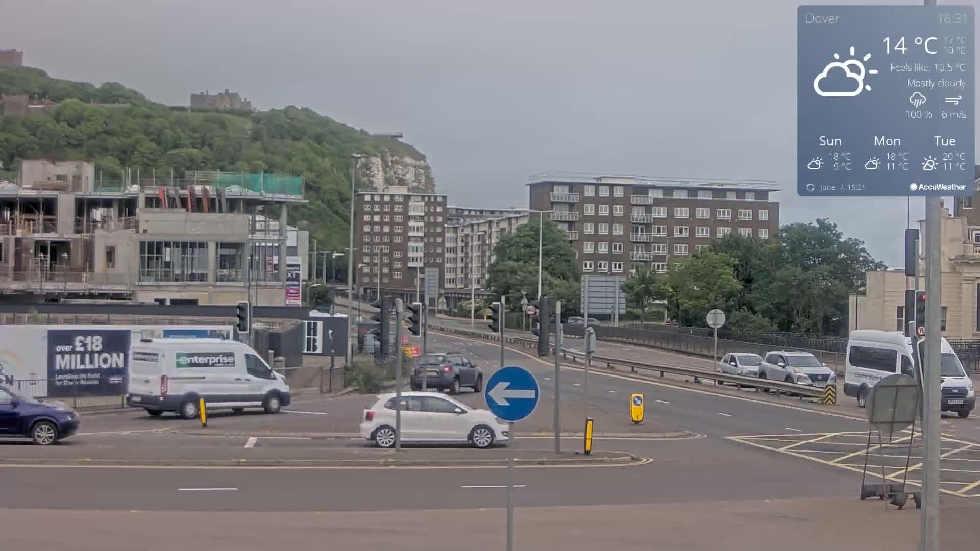 A mostly cloudy day shows a roundabout intersection with several cars and vans, a partially completed building, and apartment buildings, all set against a backdrop of green hills and white cliffs.