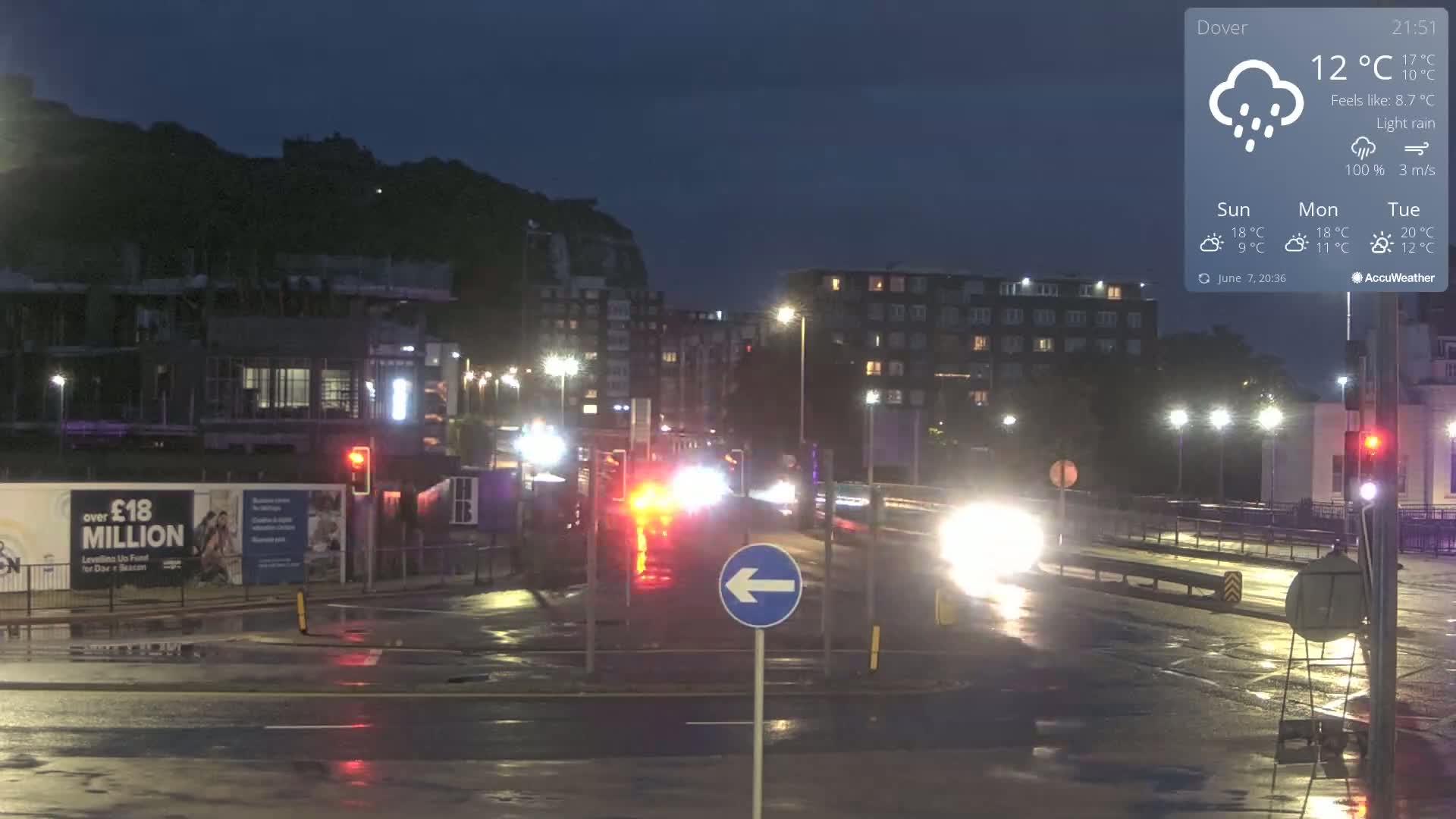 A nighttime street scene shows light rain falling on a wet road with cars driving past buildings under streetlights.