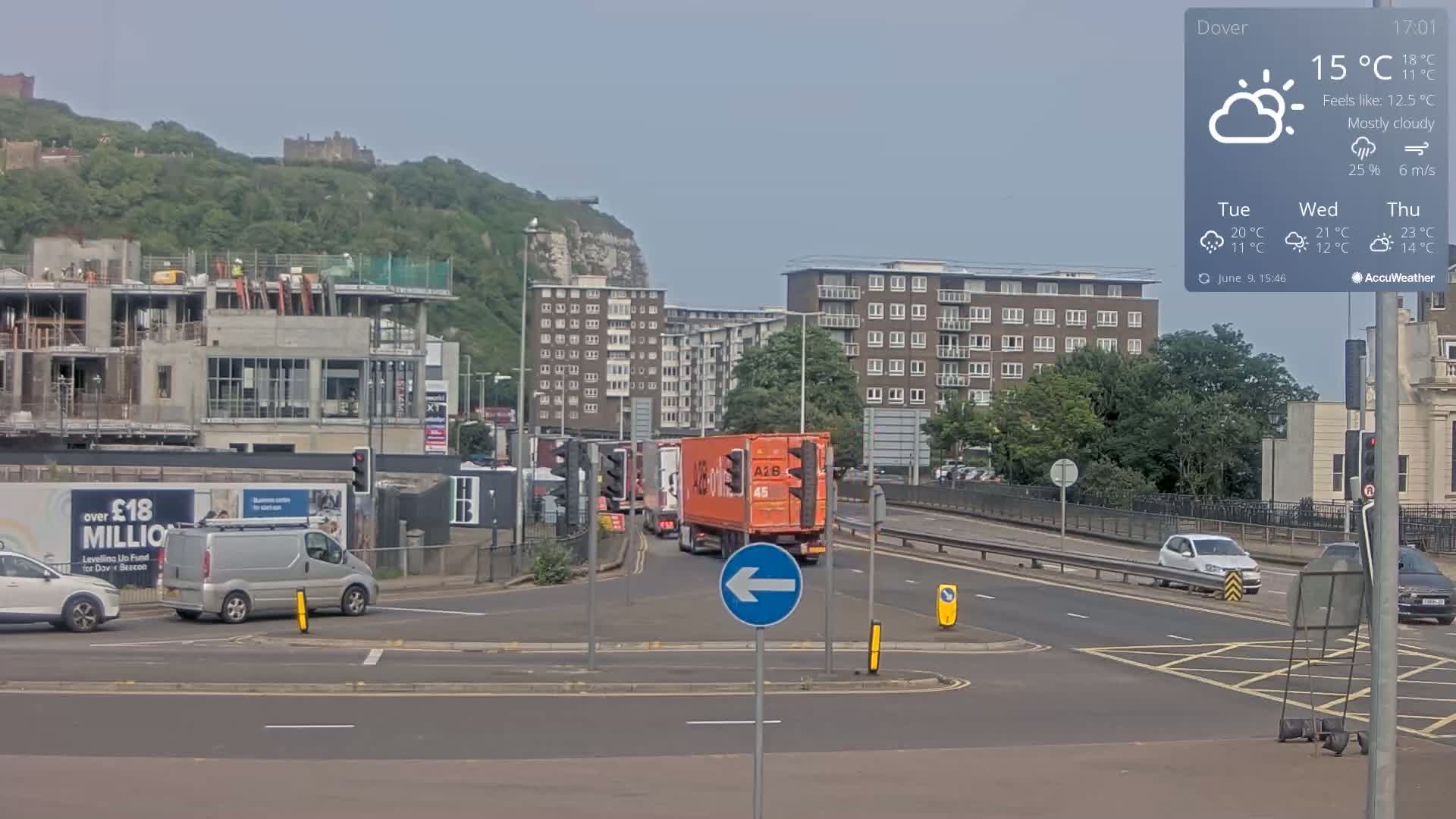 A mostly cloudy day with a temperature of 15°C shows a busy intersection with several vehicles, including a large orange semi-truck, near a construction site and residential buildings, all situated against a backdrop of a hill and a castle.