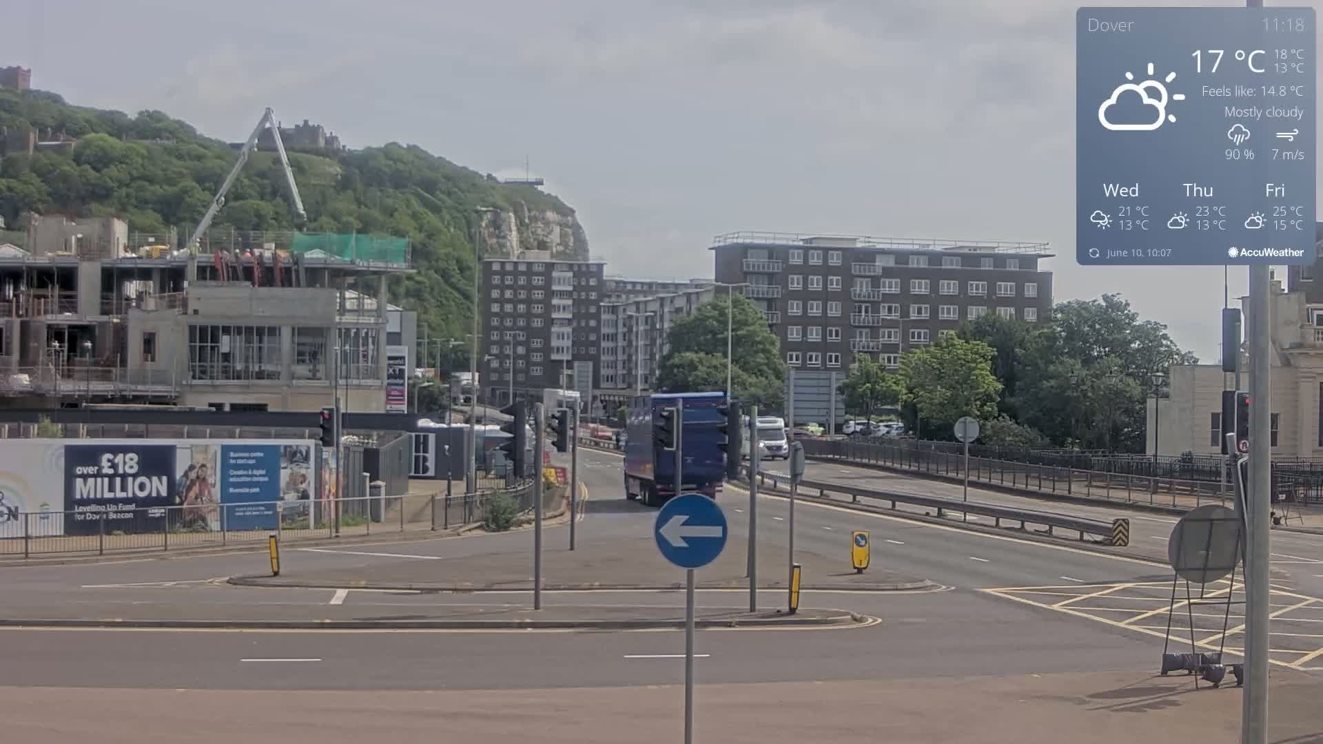 A mostly cloudy day shows a view of a road with a large construction site, apartment buildings, and a truck driving down a road, with a hill and some greenery visible in the background.