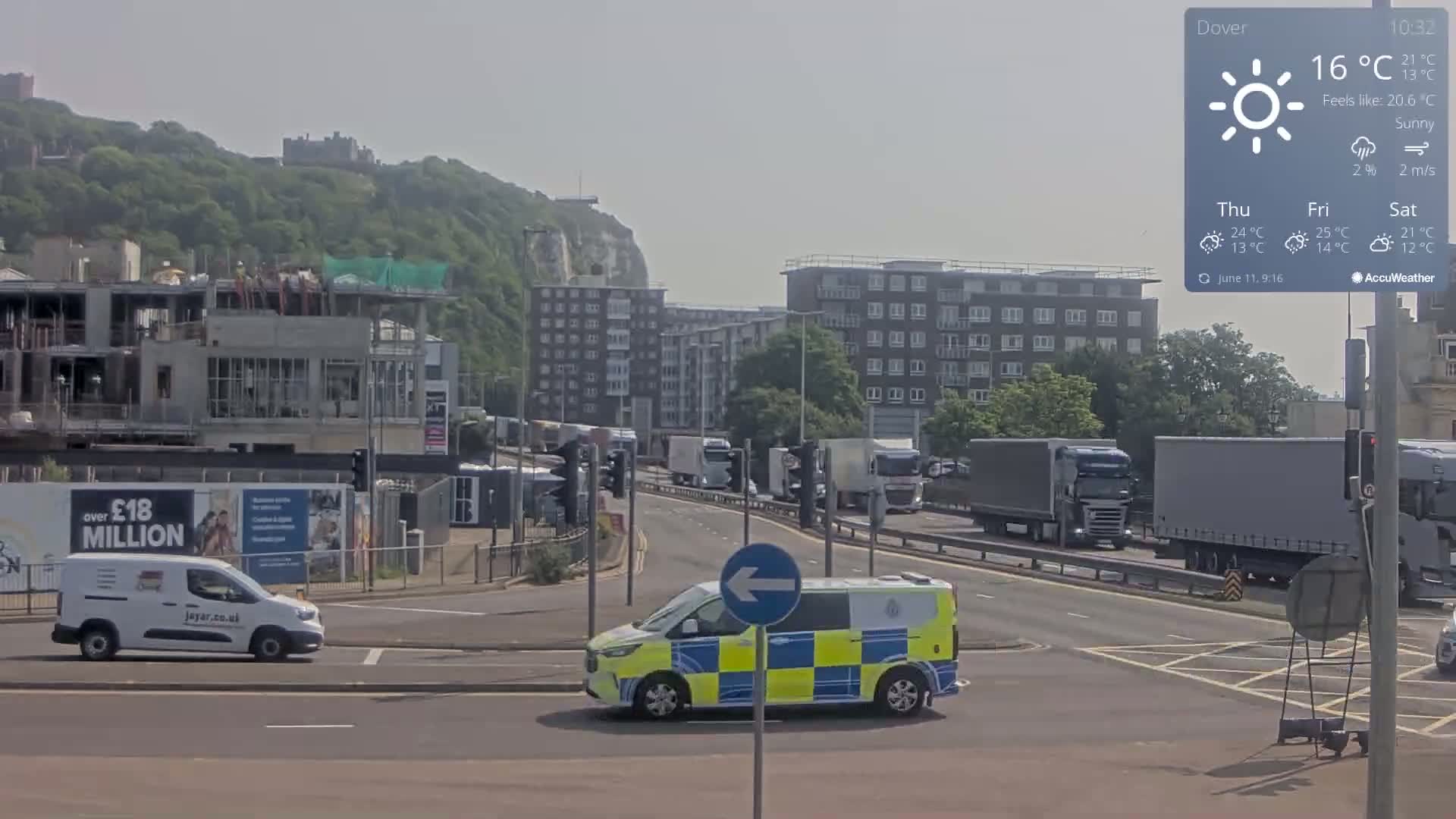 A sunny day shows a busy intersection with several vehicles, including a police van, near a construction site and apartment buildings, with white cliffs and a castle visible in the distance.