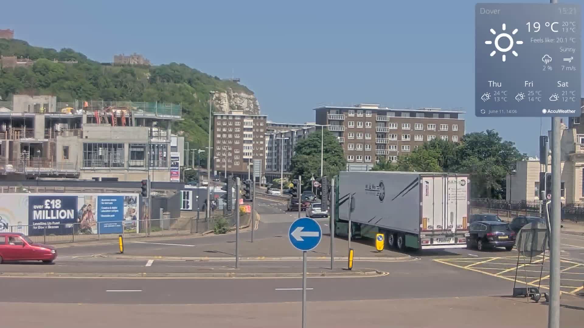 A sunny road intersection in a city with buildings, a construction site, a large white truck, and several cars, all set against a backdrop of a green hill and white cliffs.