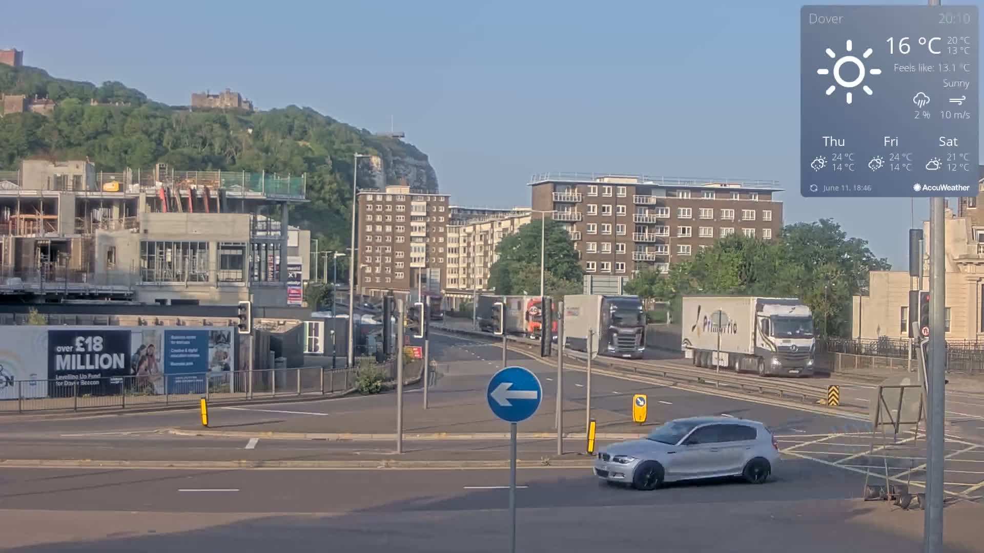 A sunny city street scene shows a silver car turning left at an intersection, with several large trucks and a building under construction in the background, all set against a backdrop of hills and castles under a sunny sky.
