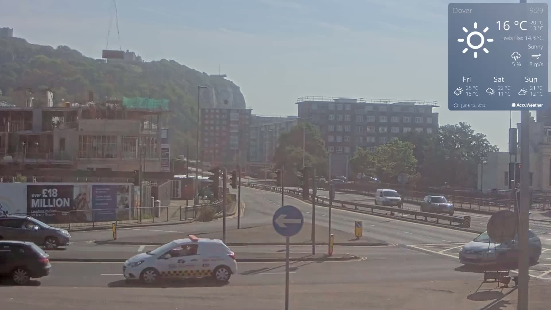 A sunny, hazy day reveals a multi-lane road with several cars, a construction site, and apartment buildings, with a large cliff and castle visible in the distance.