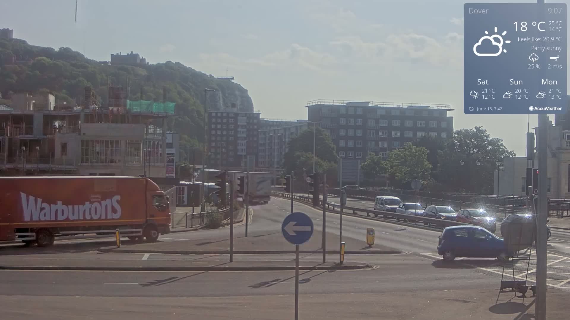 A partly sunny day shows a street scene with several cars and a large delivery truck, construction in the background and a hill with a castle visible in the distance.