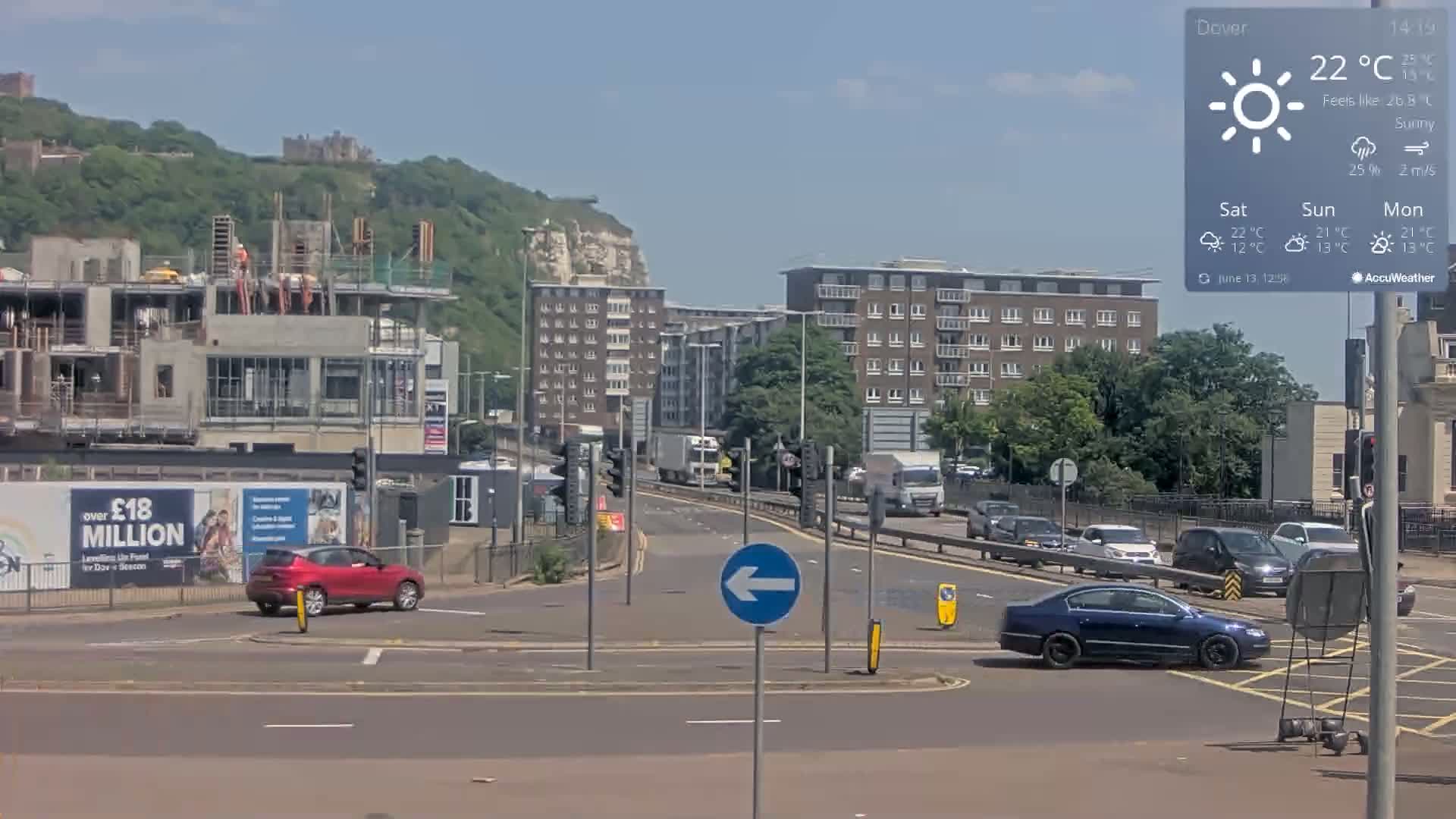 A sunny day reveals a highway intersection with several cars, a building under construction, apartment buildings, and a hill with a castle in the background.