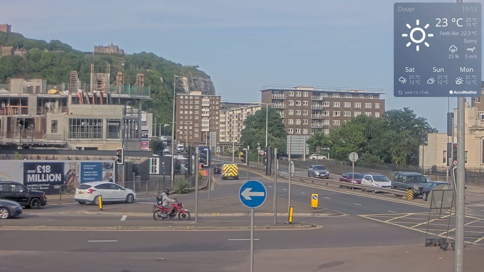 A sunny, traffic-filled road scene shows a partially constructed building, apartment buildings, a hill with a castle in the distance, and several cars and a motorcycle on the road.