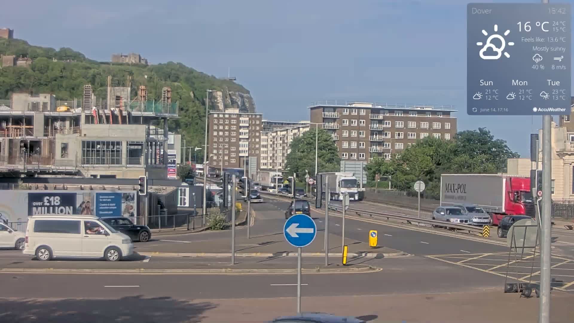 A mostly sunny, partly cloudy day shows a multi-lane road intersection with several vehicles, construction, and apartment buildings; a hill with greenery and a castle is visible in the background.