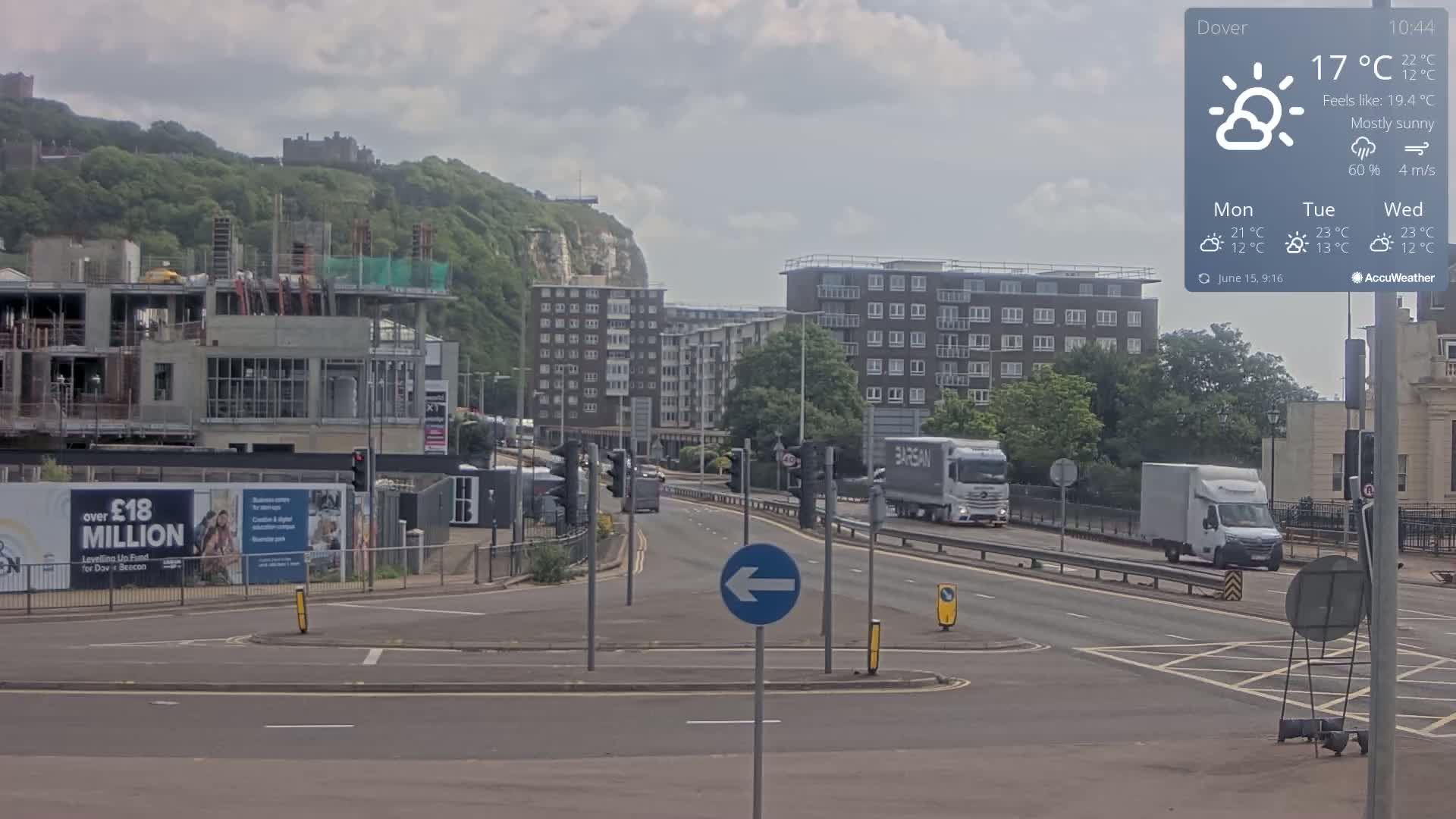 A mostly sunny, 17°C day shows a view of a multi-lane road with several vehicles, a construction site, apartment buildings, and a castle atop a green hill in the distance.