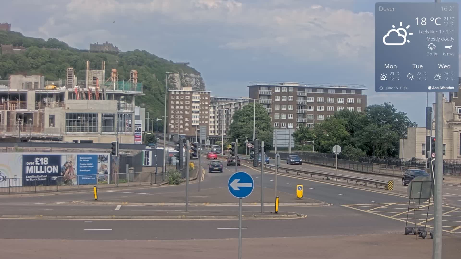 A mostly cloudy day shows a multi-lane road with several cars, a building under construction, apartment buildings, and a castle in the background on a hill.
