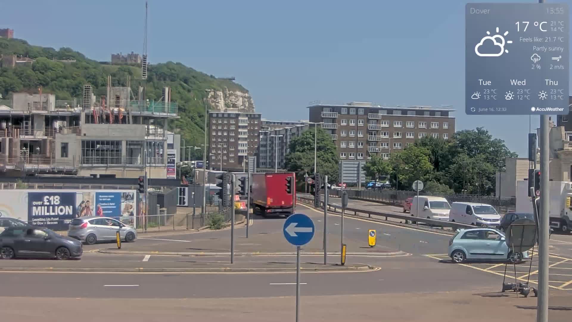 A partly sunny scene shows a multi-lane road intersection with several vehicles, a construction site, apartment buildings, and a distant cliffside.