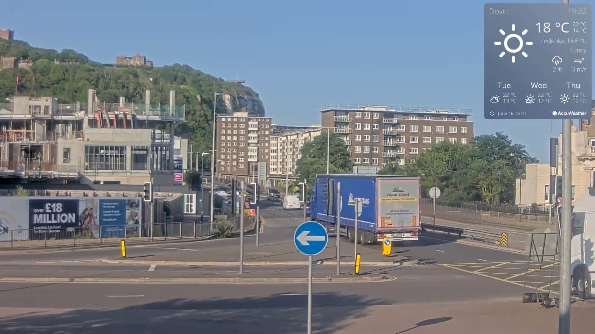 A sunny day shows a partially constructed building, several apartment buildings, and a large blue semi-truck at a roundabout, with a hill and castle visible in the background.