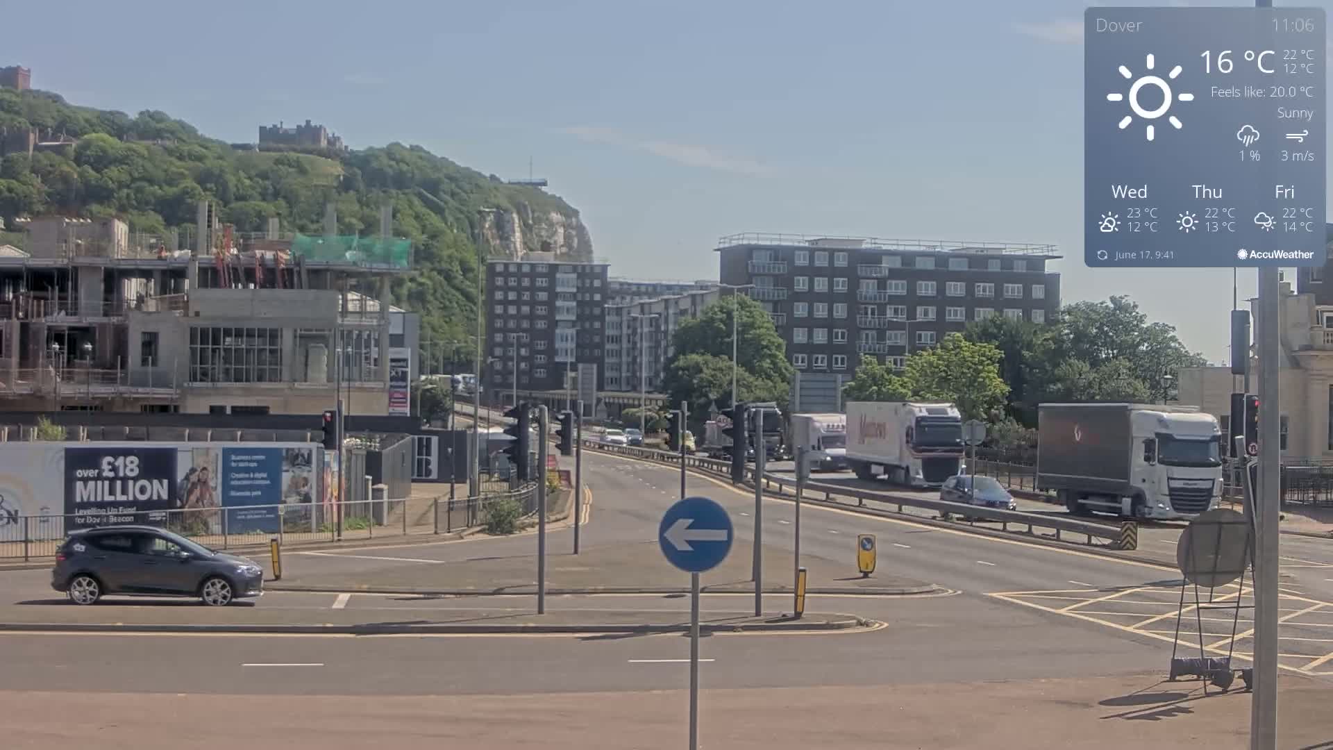 A sunny day shows a multi-lane road with cars and trucks, a construction site, and apartment buildings, all set against a backdrop of green hills and white cliffs.