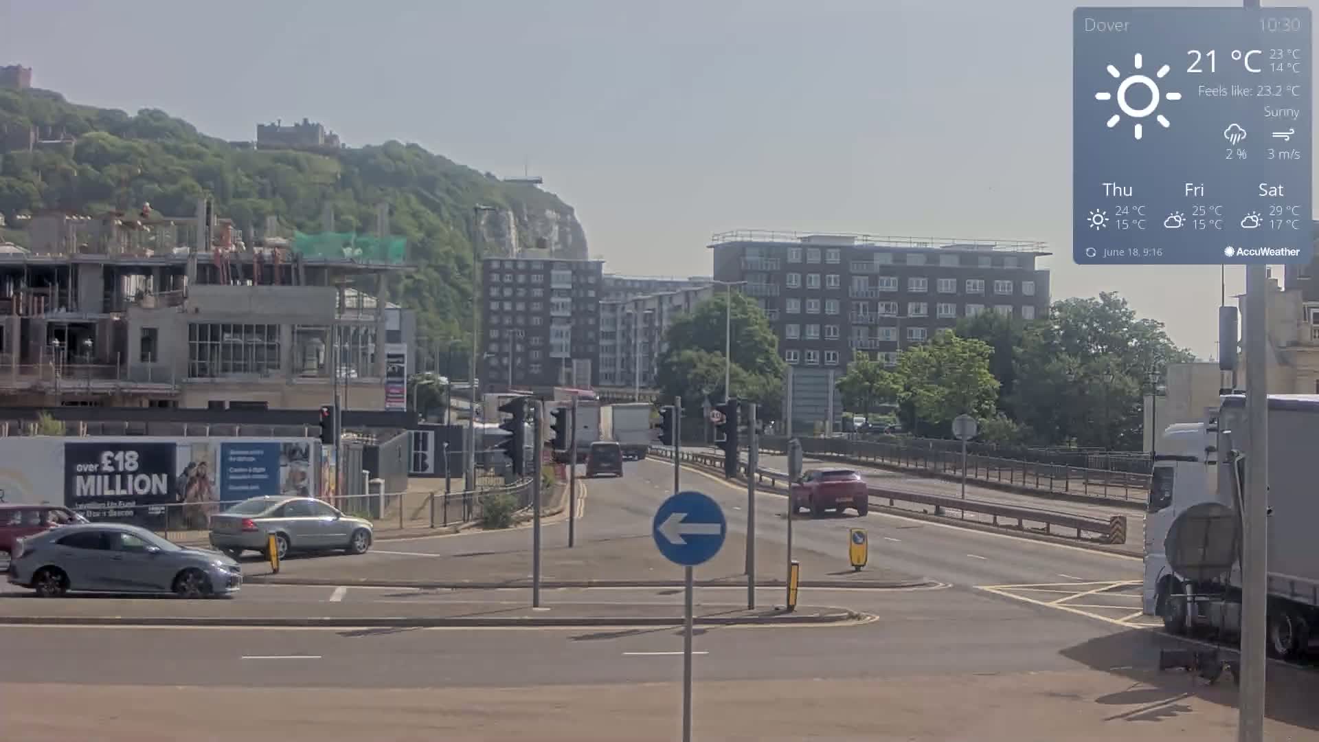 A sunny, 21°C day shows a roadway intersection with several cars and a large construction site near a hill and white cliffs in the background.