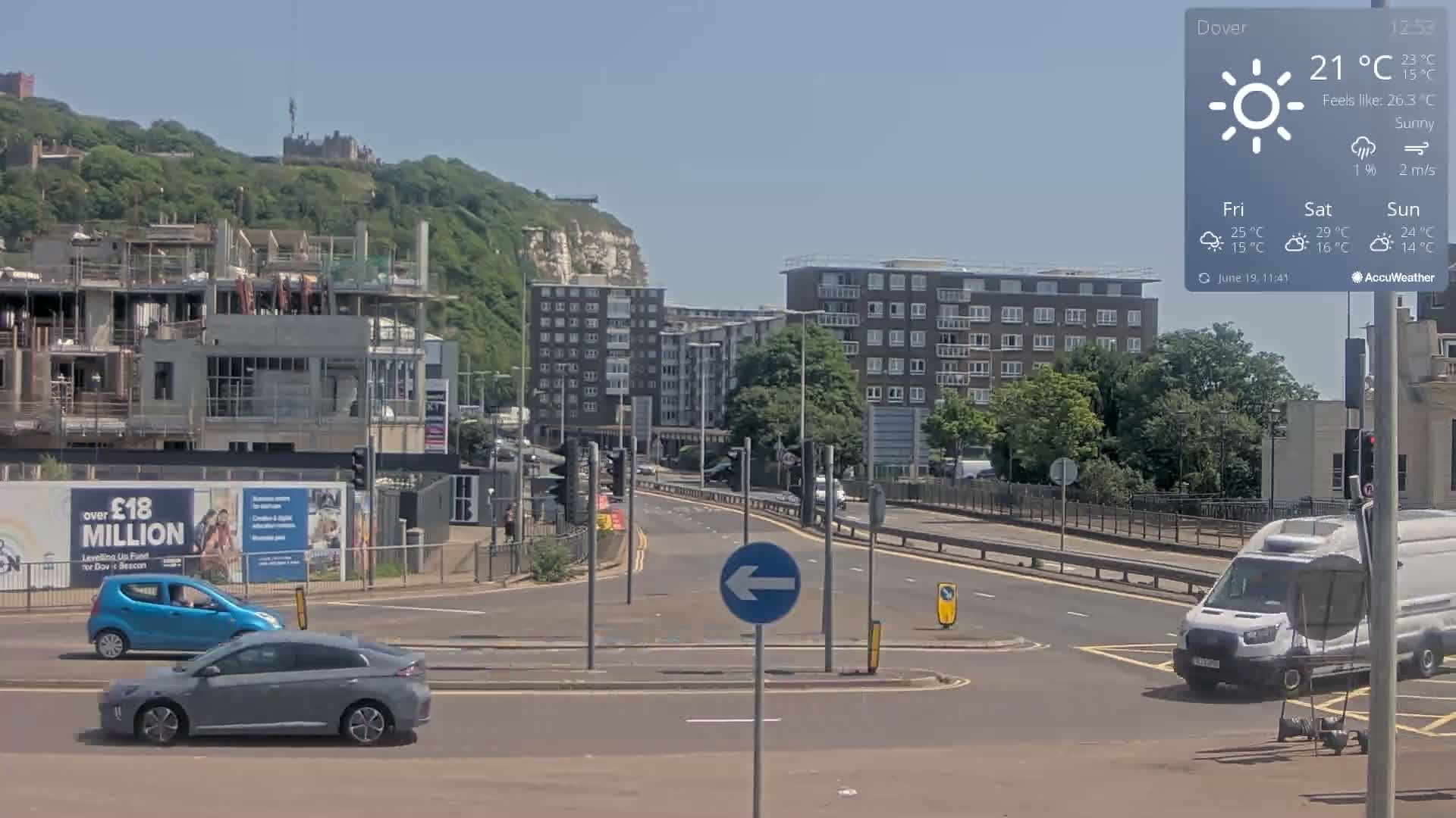 A sunny day shows a mostly empty road with a few cars and a white van, with a partly constructed building and residential buildings in the background, all situated against a backdrop of green hills and a white cliff.