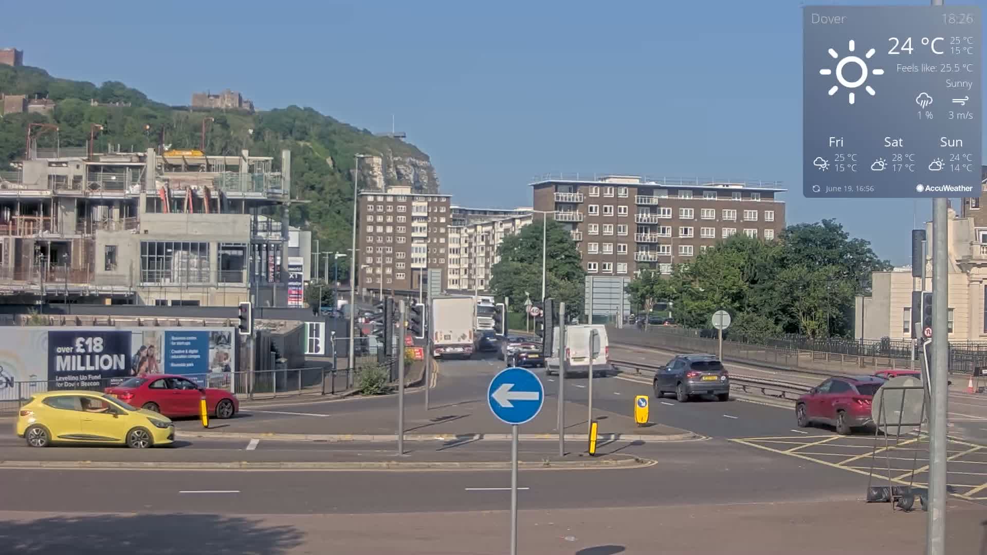 A sunny day shows a busy intersection with several cars and a large construction site, all situated near a hill with buildings and what appears to be a castle in the distance.