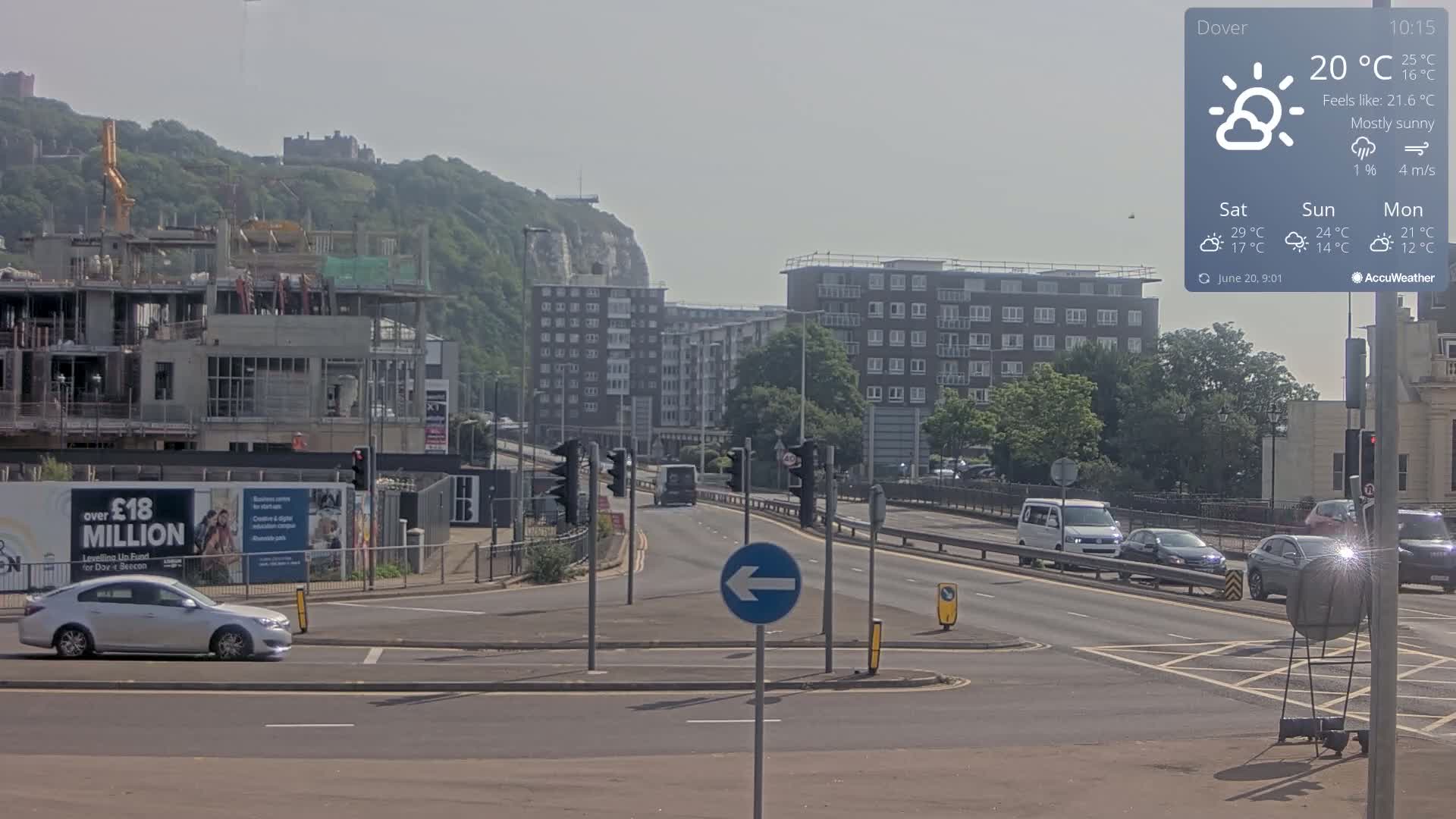 A mostly sunny day shows a multi-lane road with several cars, a construction site, and apartment buildings, all set against a backdrop of a hill with a castle and white cliffs.