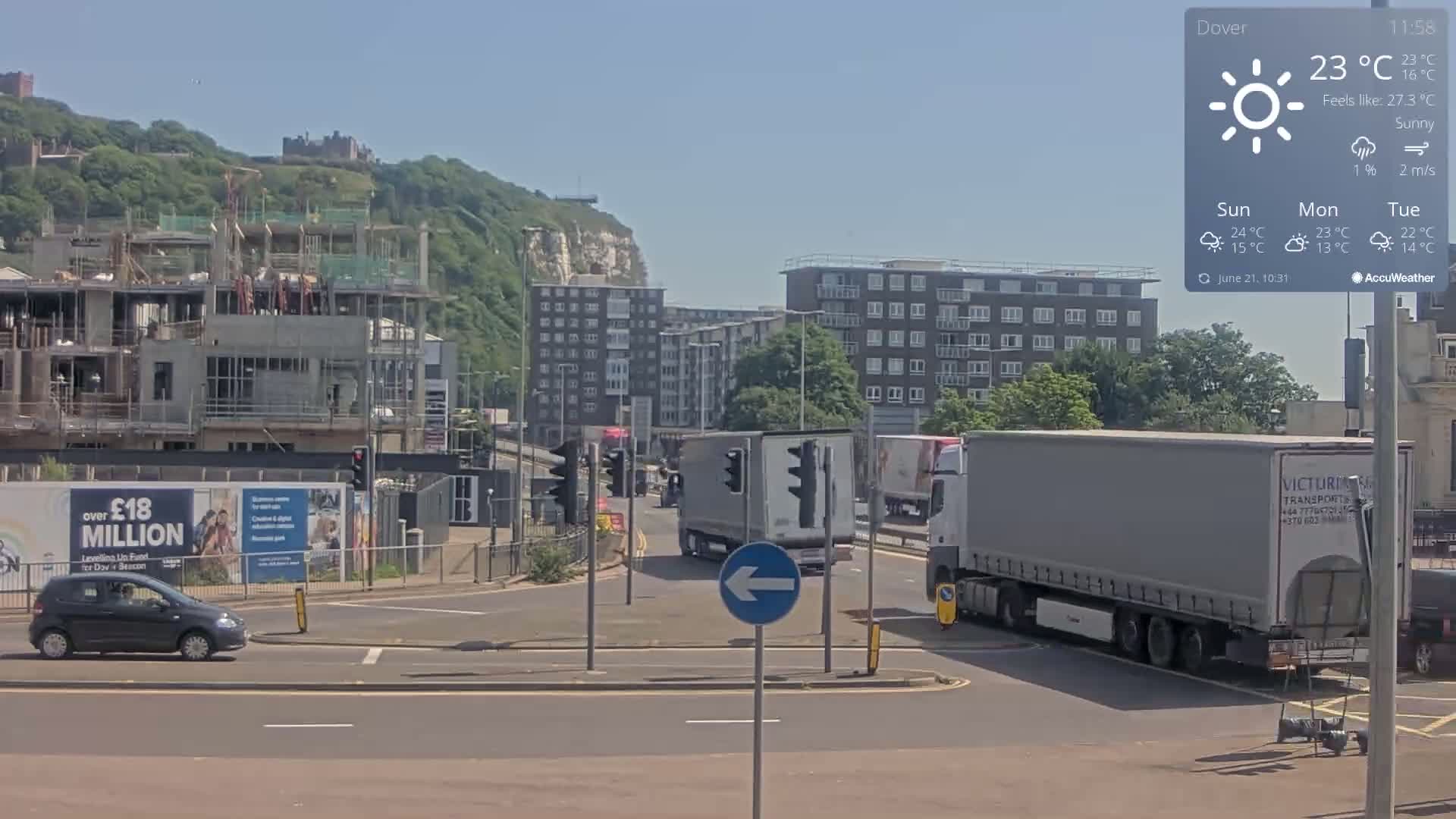 Under sunny skies, a busy roadway intersects near a construction site and apartment buildings, with a hill and castle in the background.