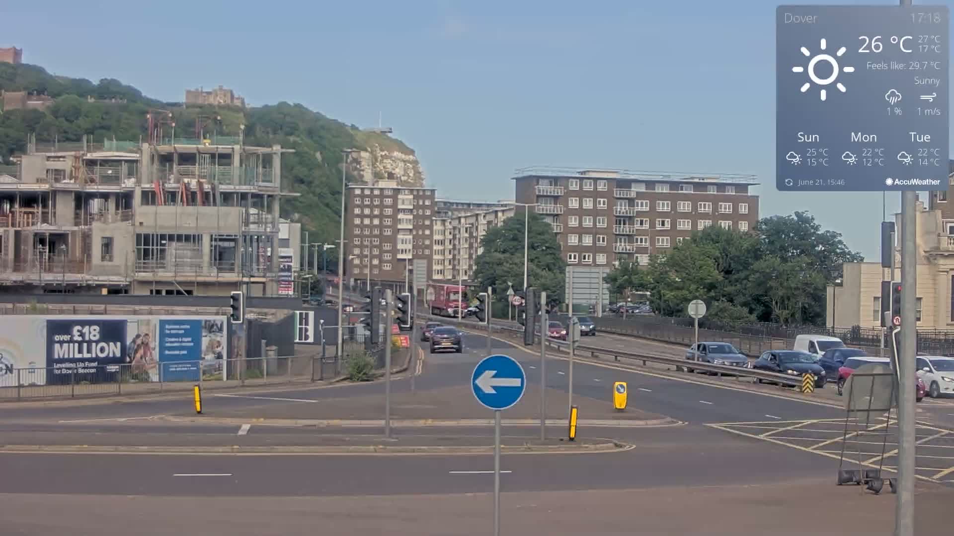 A sunny day shows a busy intersection with several cars, a partially constructed building, and apartment buildings, all set against a backdrop of a hill with a castle and cliffs.