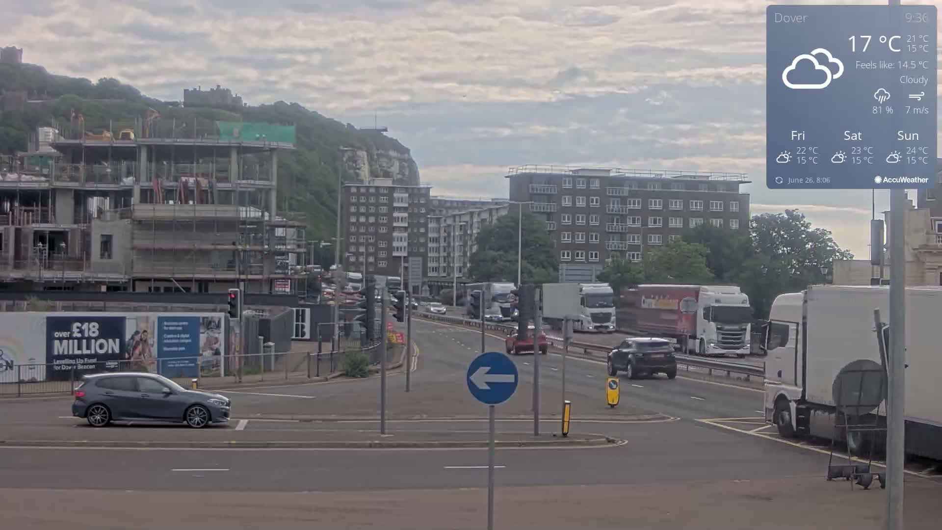 A cloudy day shows a busy roadway with several vehicles, including trucks and cars, passing through a crossroads near a construction site and apartment buildings against a backdrop of a hill with a castle.