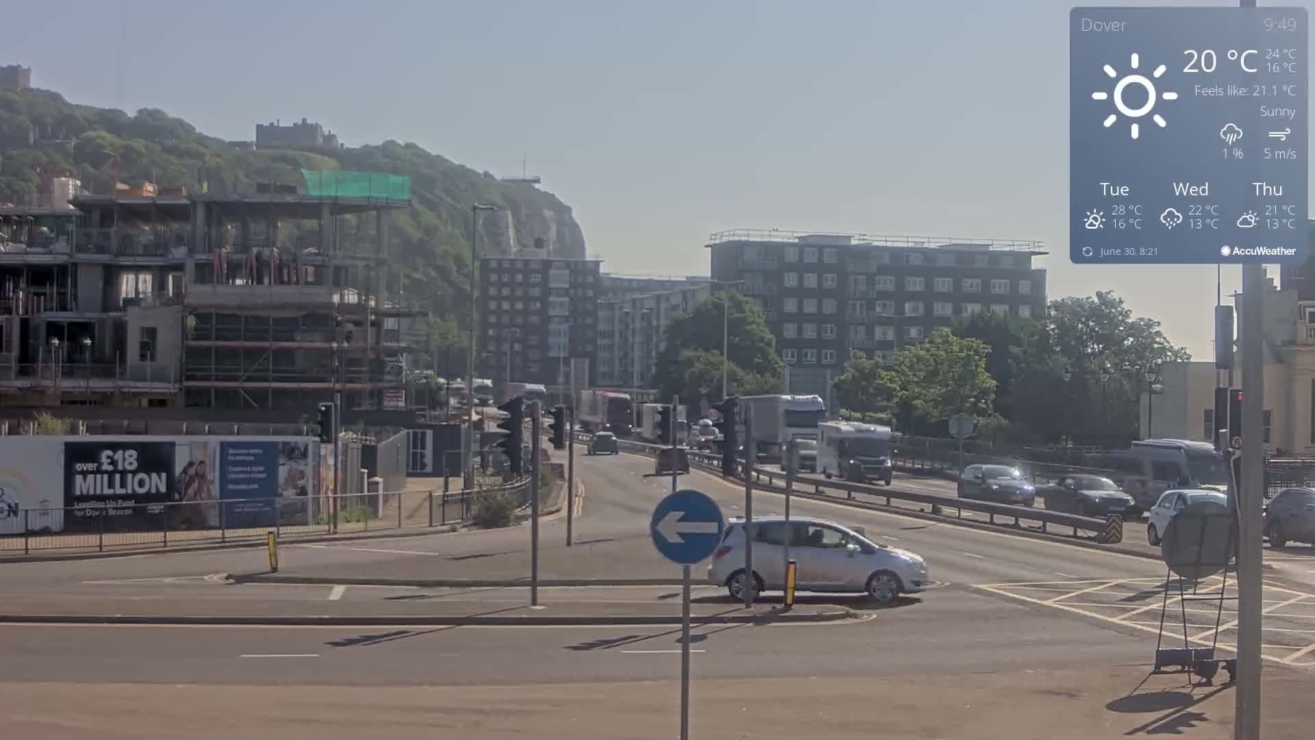 A sunny day shows a busy road intersection with cars and trucks, a partially constructed building, and a cliff in the background.
