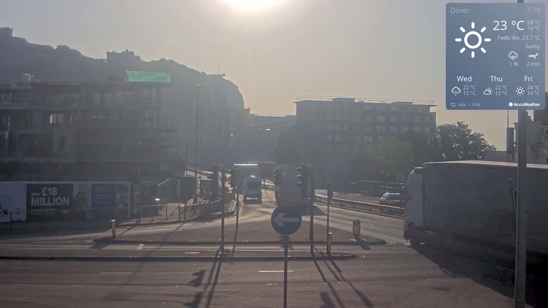 A hazy, sunny morning shows a multi-lane road with several vehicles, buildings under construction, and a distant hill with a castle in the background.
