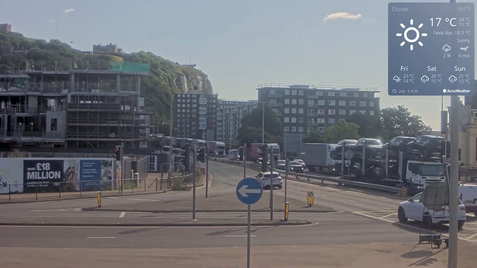 A sunny day shows a multi-lane road with moderate traffic, including a car carrier, near a construction site and apartment buildings, with a white cliff and castle visible in the distance.