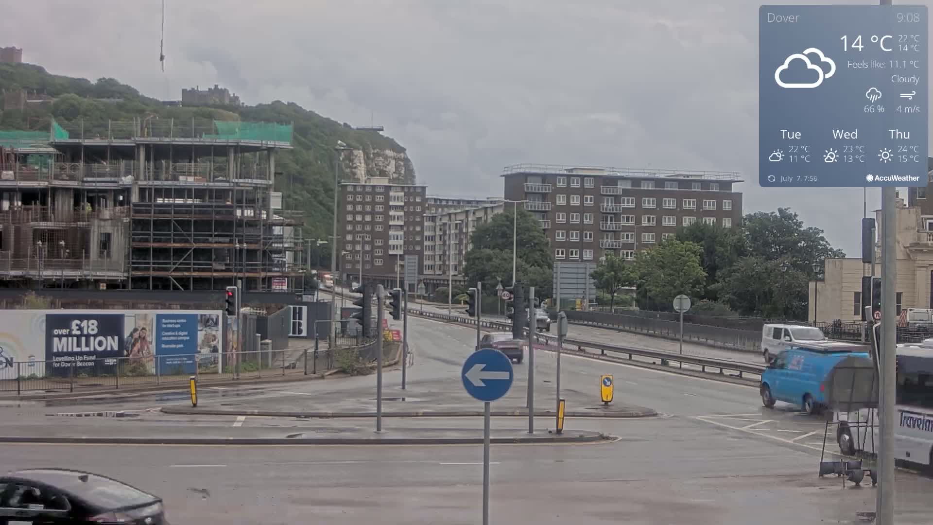 A cloudy day shows a busy intersection with several cars and vans, a partially constructed building, and apartment buildings against a backdrop of green hills and a castle.