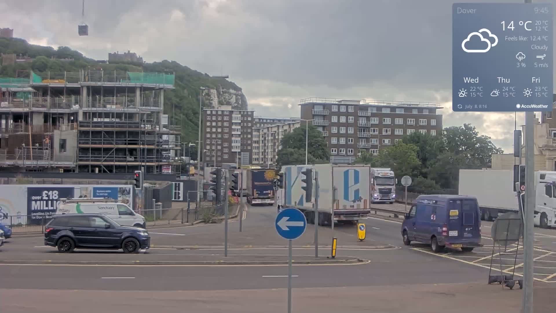A cloudy day reveals a busy intersection with several vehicles, including trucks and vans, near a large construction site and apartment buildings, all set against a backdrop of green hills and a castle.