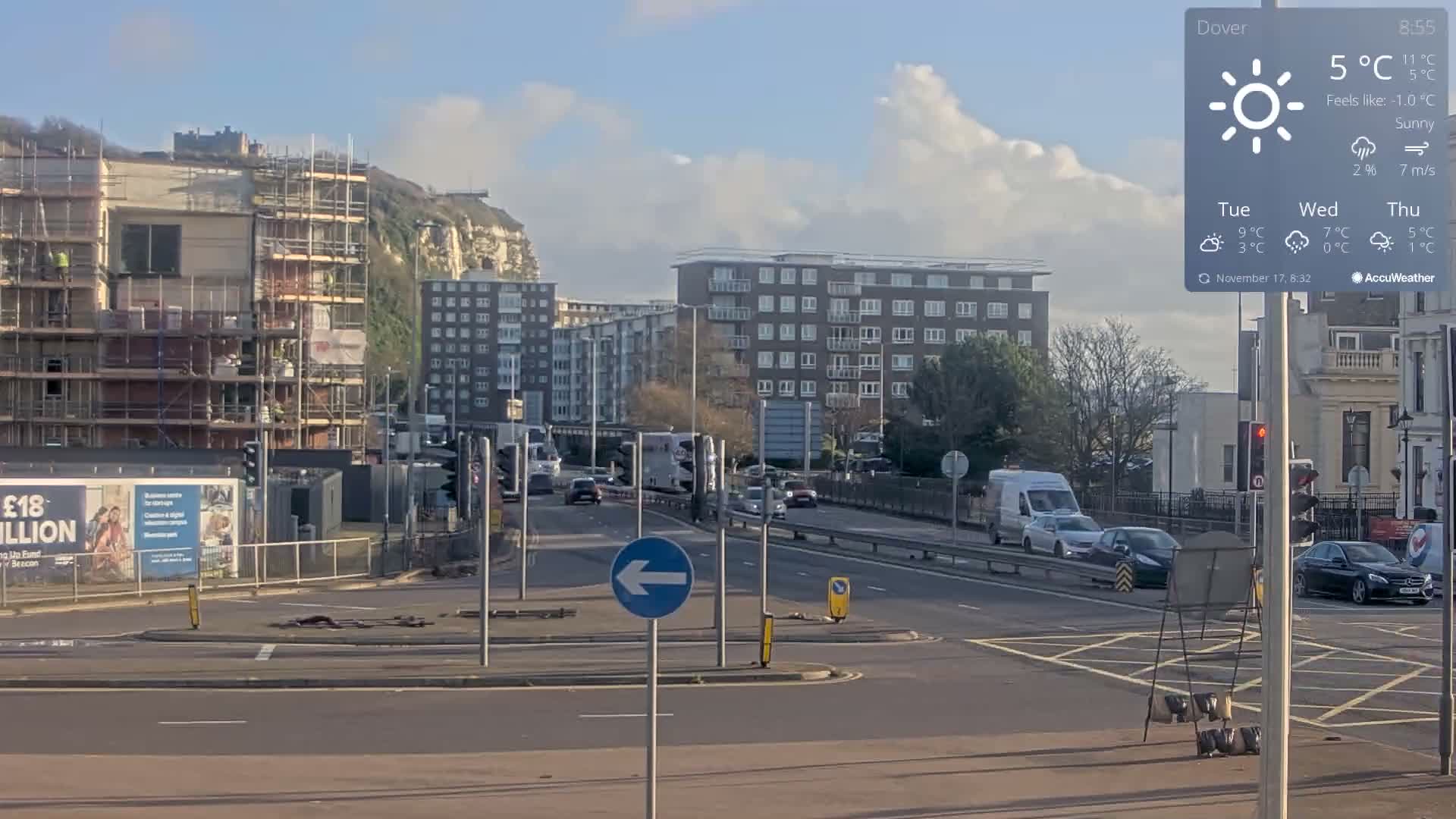 An urban street scene on a sunny day features a multi-lane road with cars and traffic lights, flanked by multi-story buildings, including one under construction with scaffolding, all beneath a partially cloudy sky and overlooked by a prominent white cliff in the background, with the air feeling crisp at around 5°C.