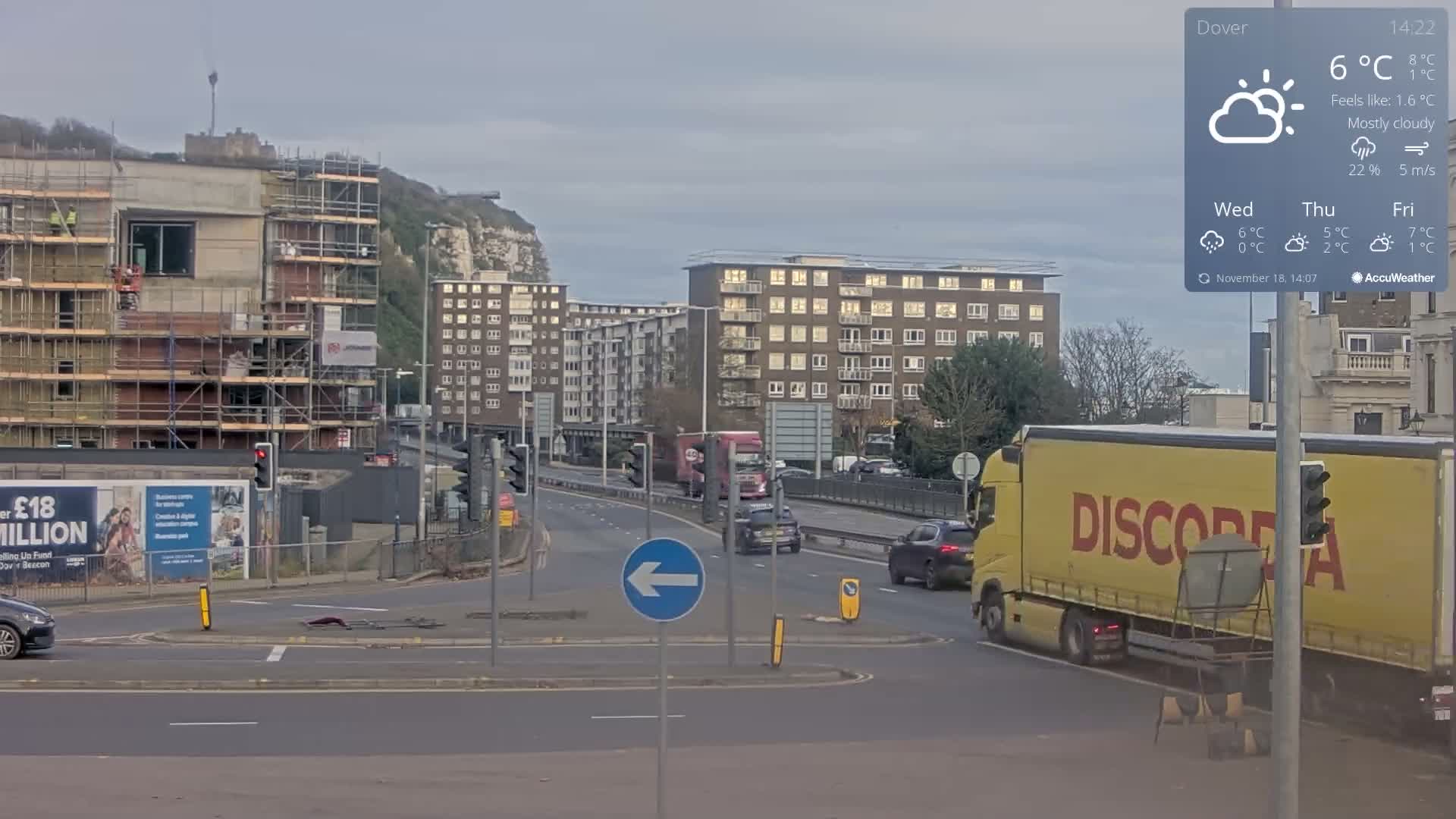 An urban street scene on a sunny day features a multi-lane road with cars and traffic lights, flanked by multi-story buildings, including one under construction with scaffolding, all beneath a partially cloudy sky and overlooked by a prominent white cliff in the background, with the air feeling crisp at around 5°C.