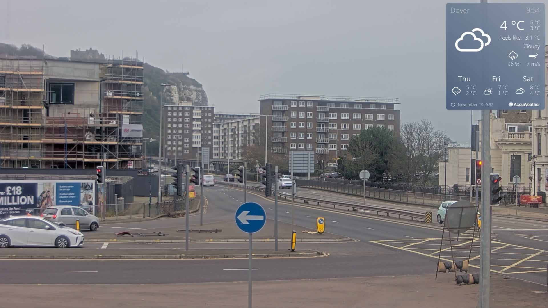 A cloudy day overlooks a bustling urban road junction with ongoing construction, multiple cars, and distant white cliffs, surrounded by buildings and leafless trees.